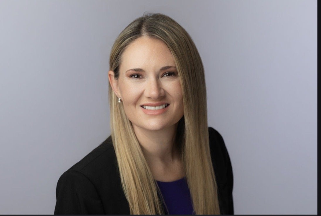 A woman with long blonde hair, smiling, in professional attire, against a light gray background.
