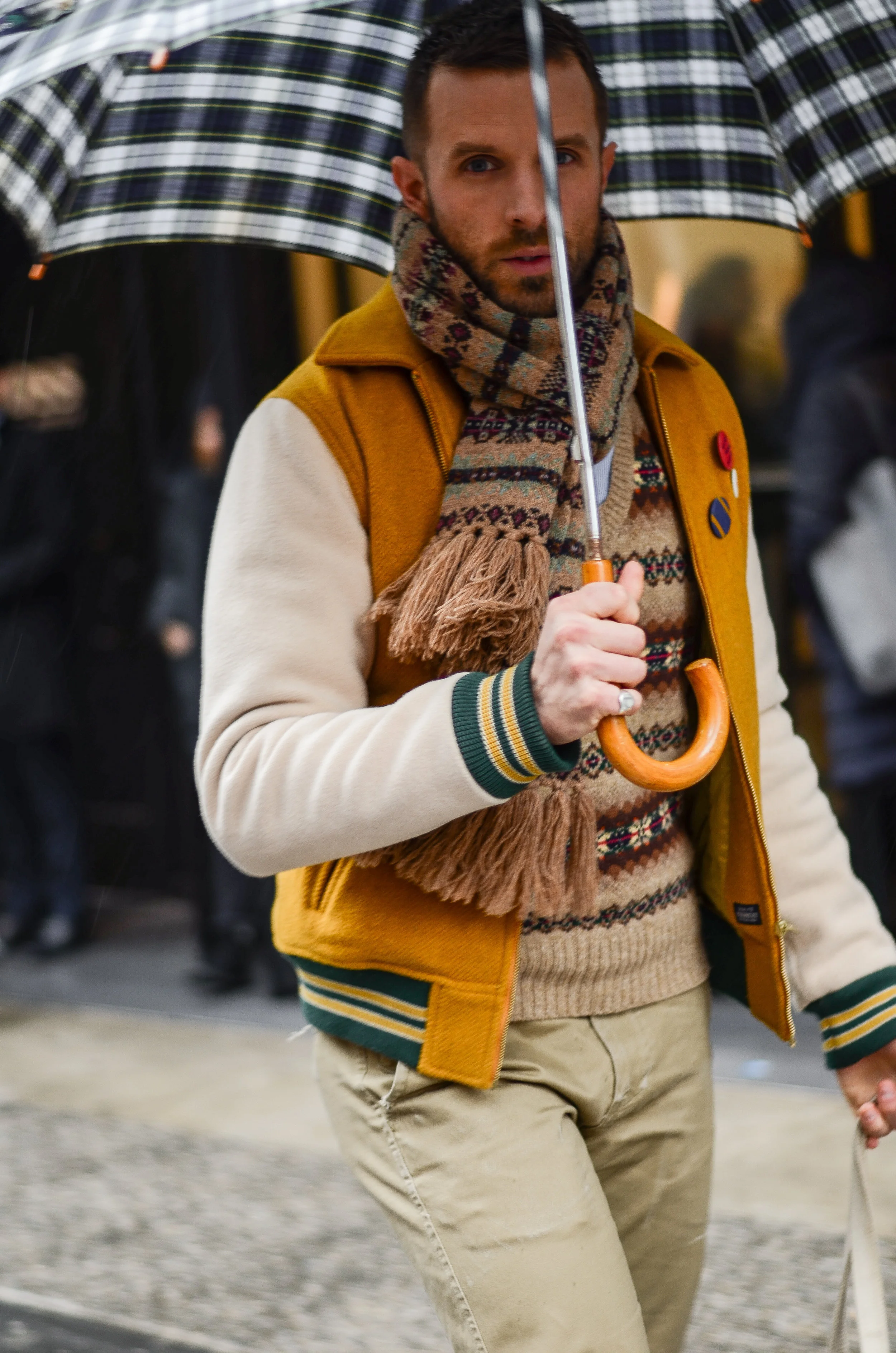 His camel varsity jacket provides a youthful foundation for his fair-isle knit sweater and scarf, and plaid umbrella.