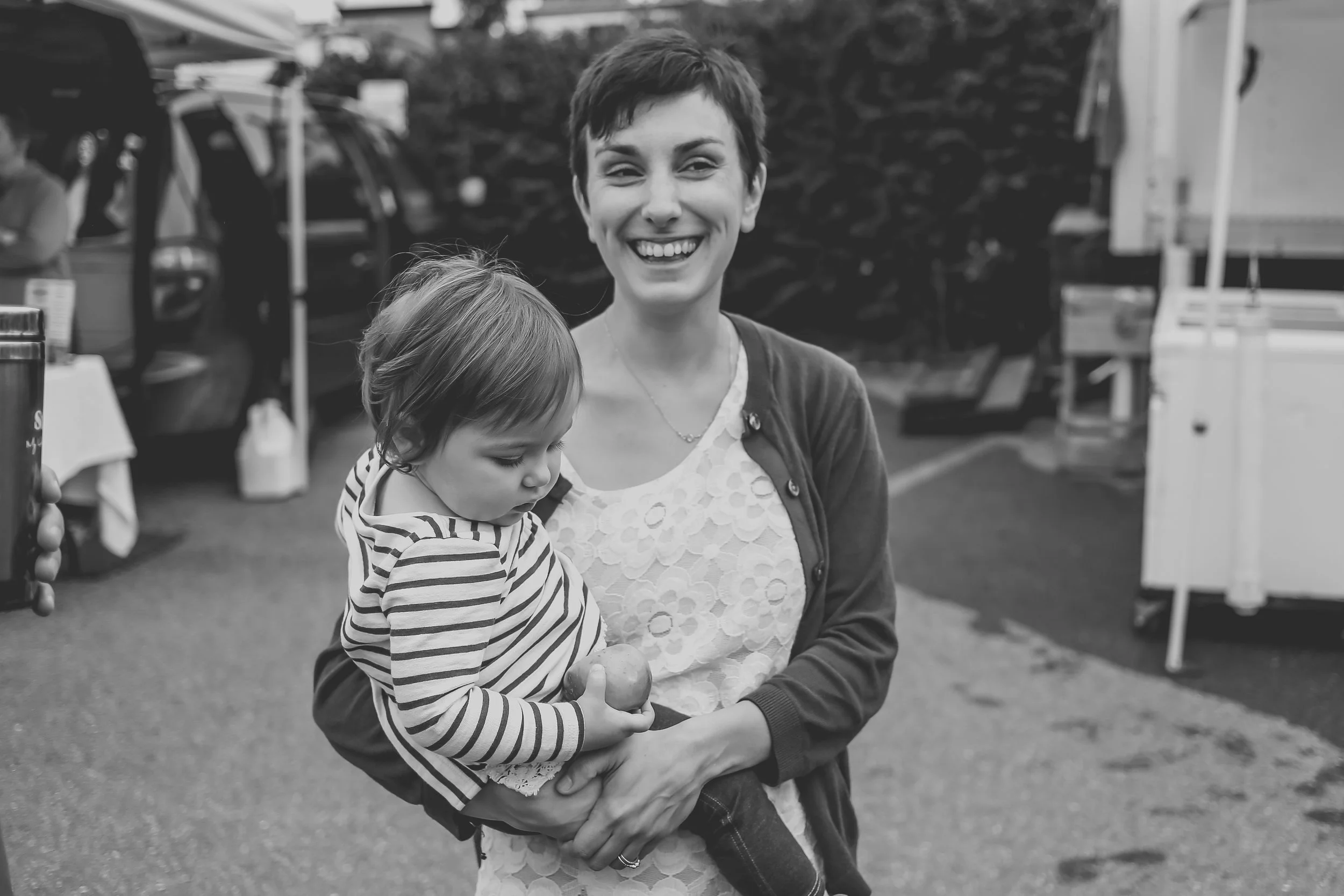 Yound mother smiling while holding her toddler daughter at the Emmaus Farmer's Market in Lehigh Valley, PA. Pennsylvania documentary family photography.