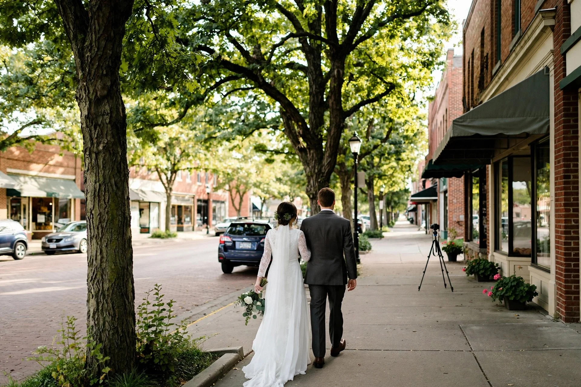 Bride and groom walking under mature oak trees near a brick downtown street in Downers Grove, Illinois, with a small DSLR camera on a tripod filming discreetly