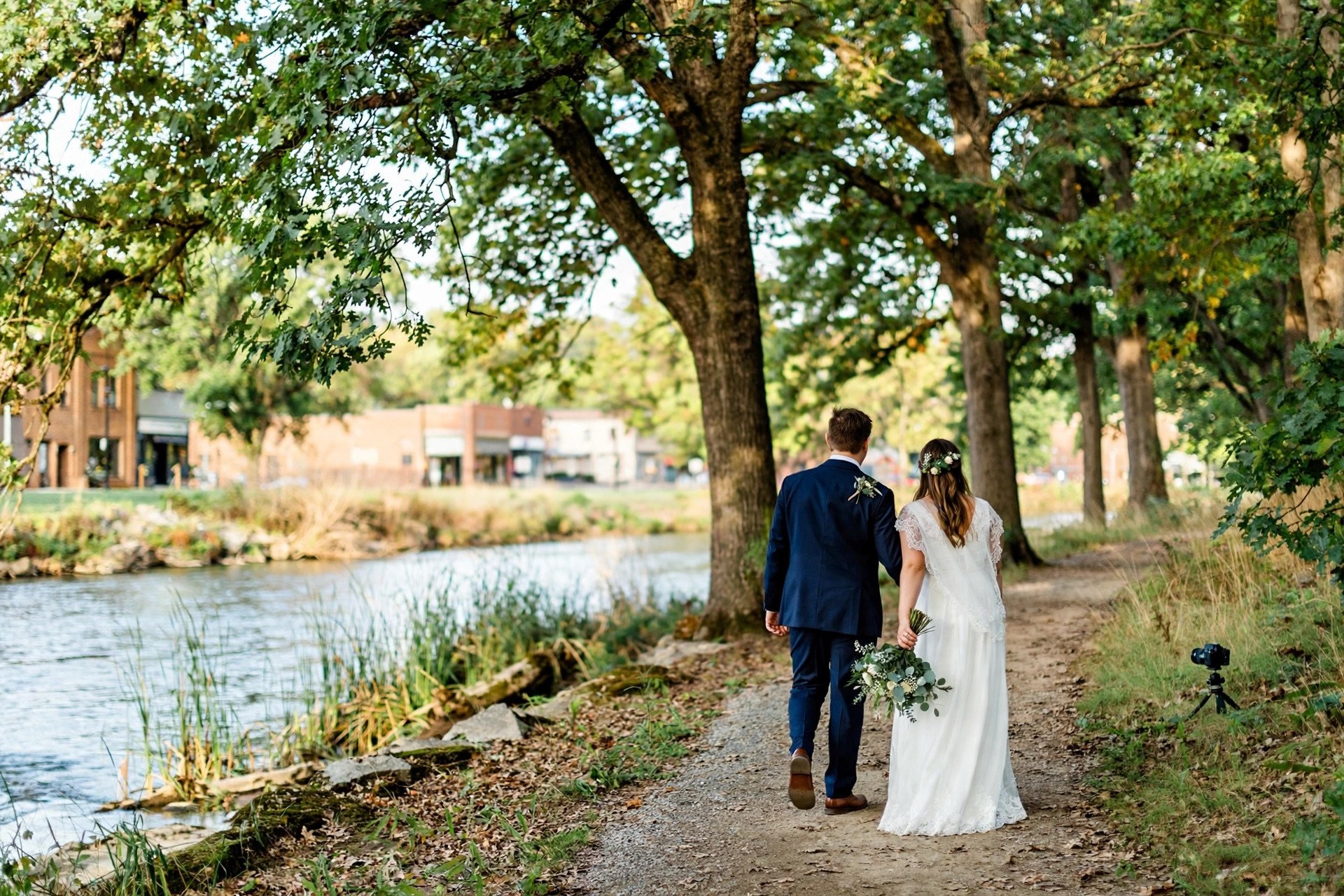 Bride and groom walking along the Des Plaines River trail under tall oak trees in Des Plaines, Illinois, with a small DSLR camera on a tripod filming discreetly nearby