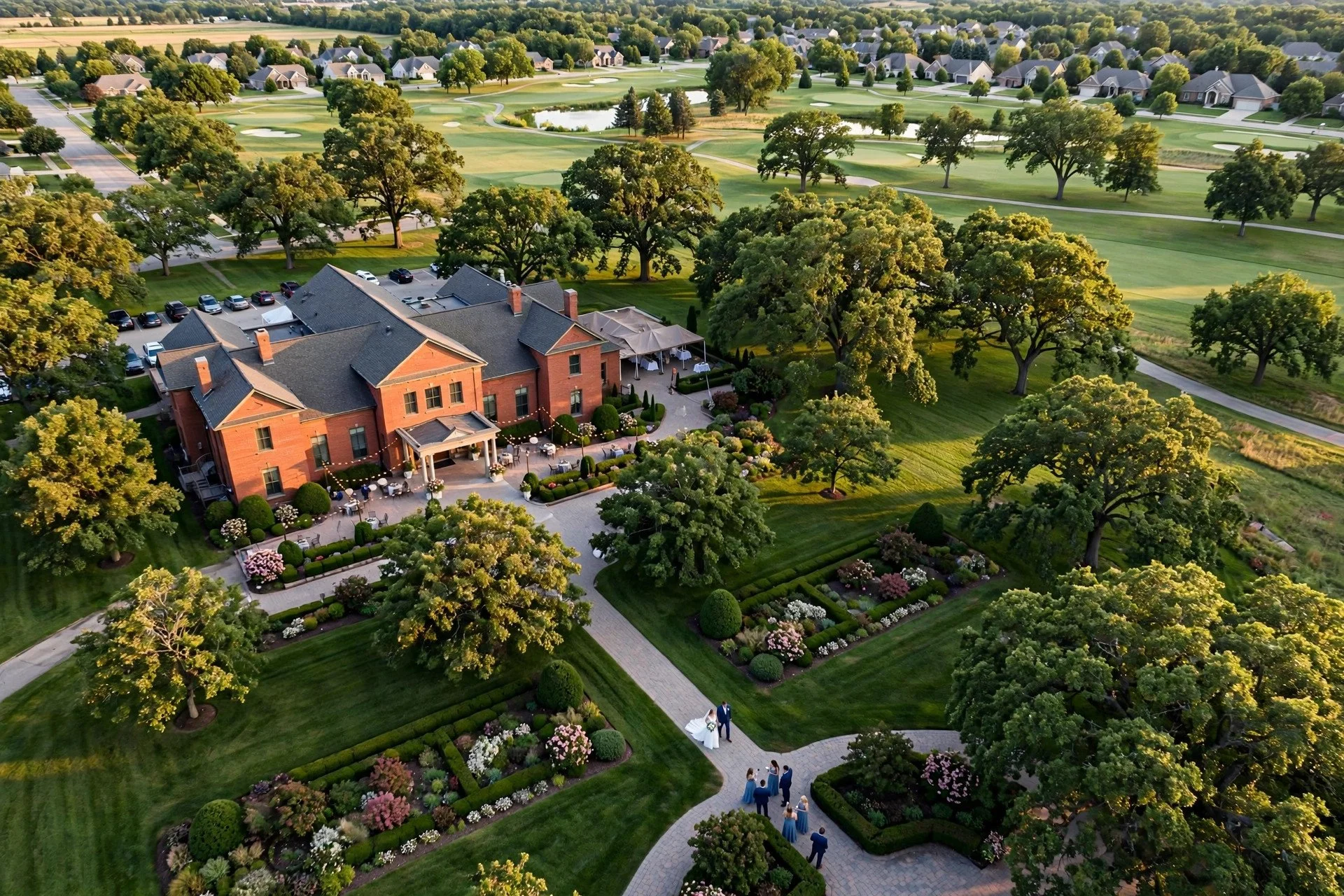 Aerial view of a suburban Midwest wedding venue with a brick ballroom, green lawn, and oak trees in Illinois, bride and groom walking together below as a small drone captures the scene from above.
