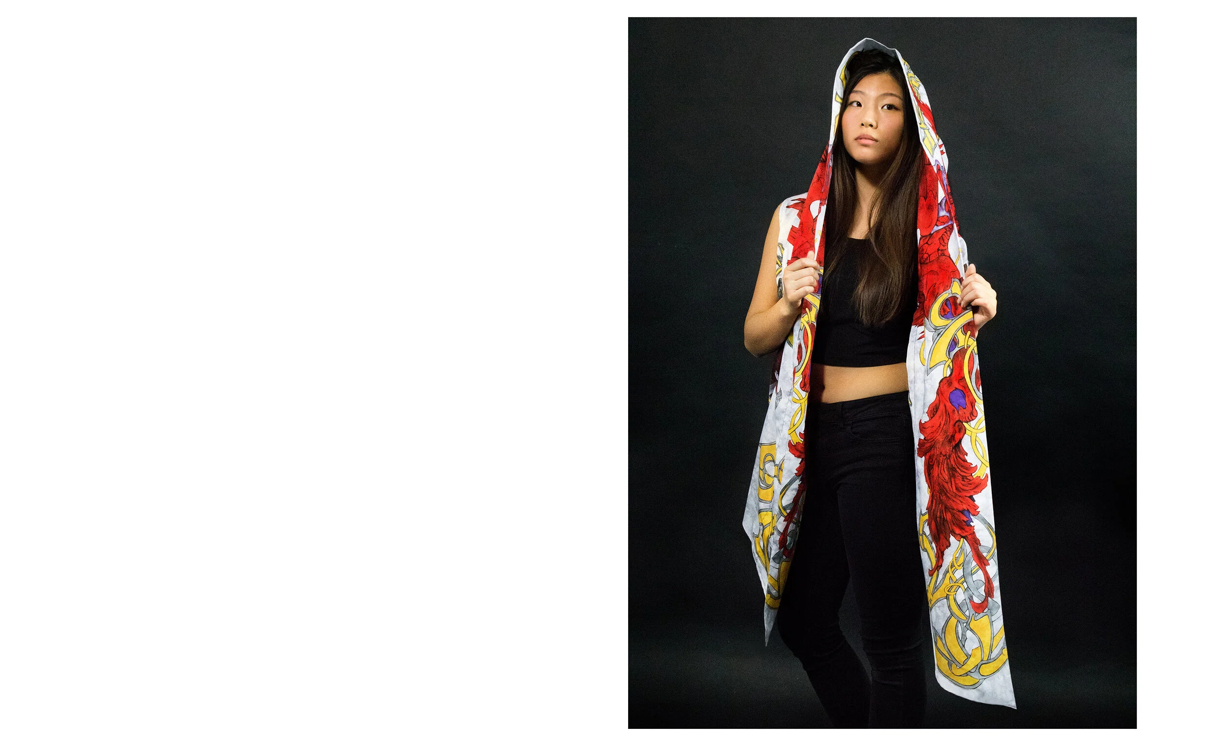 Young woman with long dark hair wearing a black crop top and black pants, holding a colorful patterned long scarf with both hands, standing against a black background.