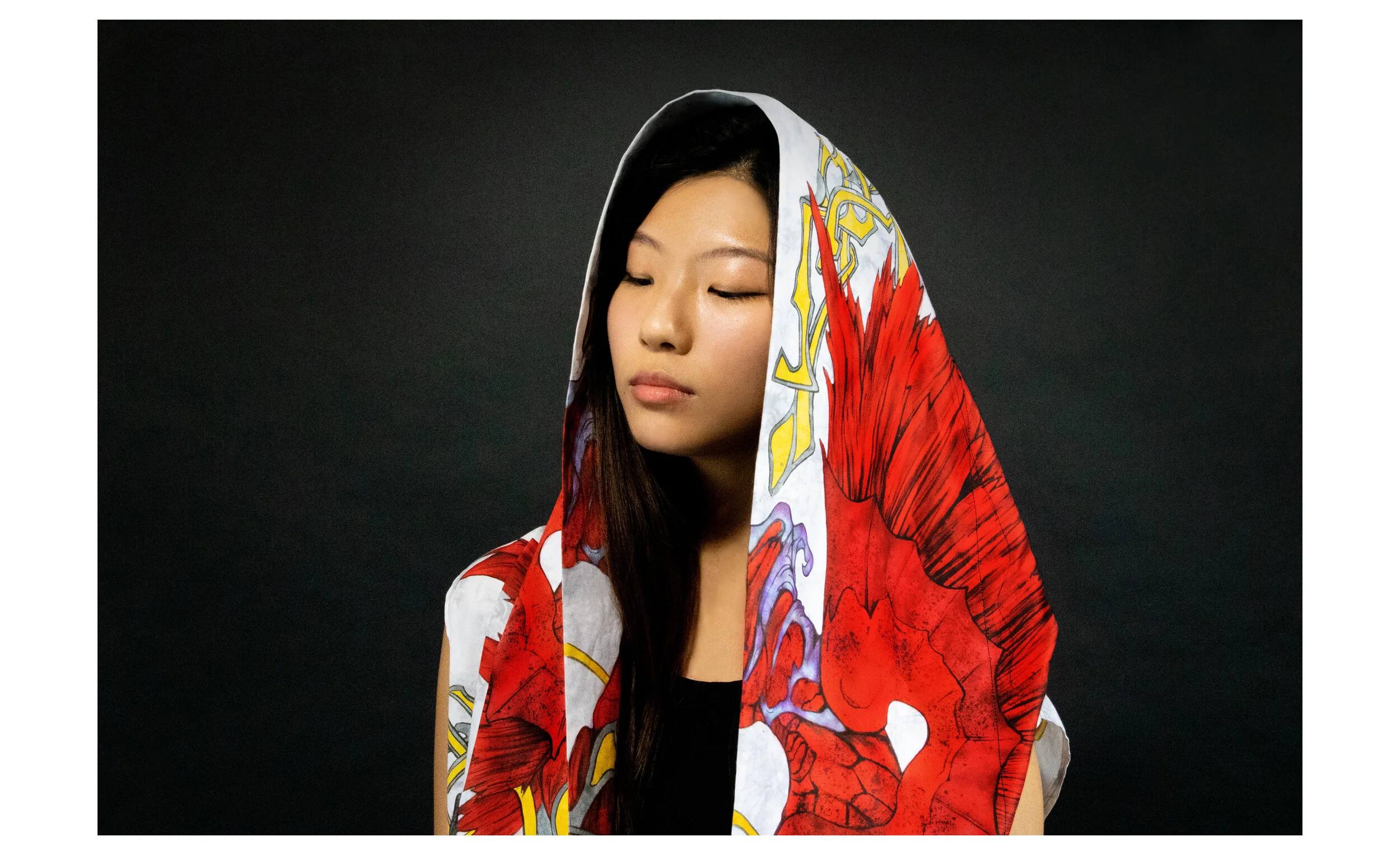 A young woman with long dark hair, closed eyes, and a peaceful expression, wearing a colorful, patterned hooded garment against a black background.