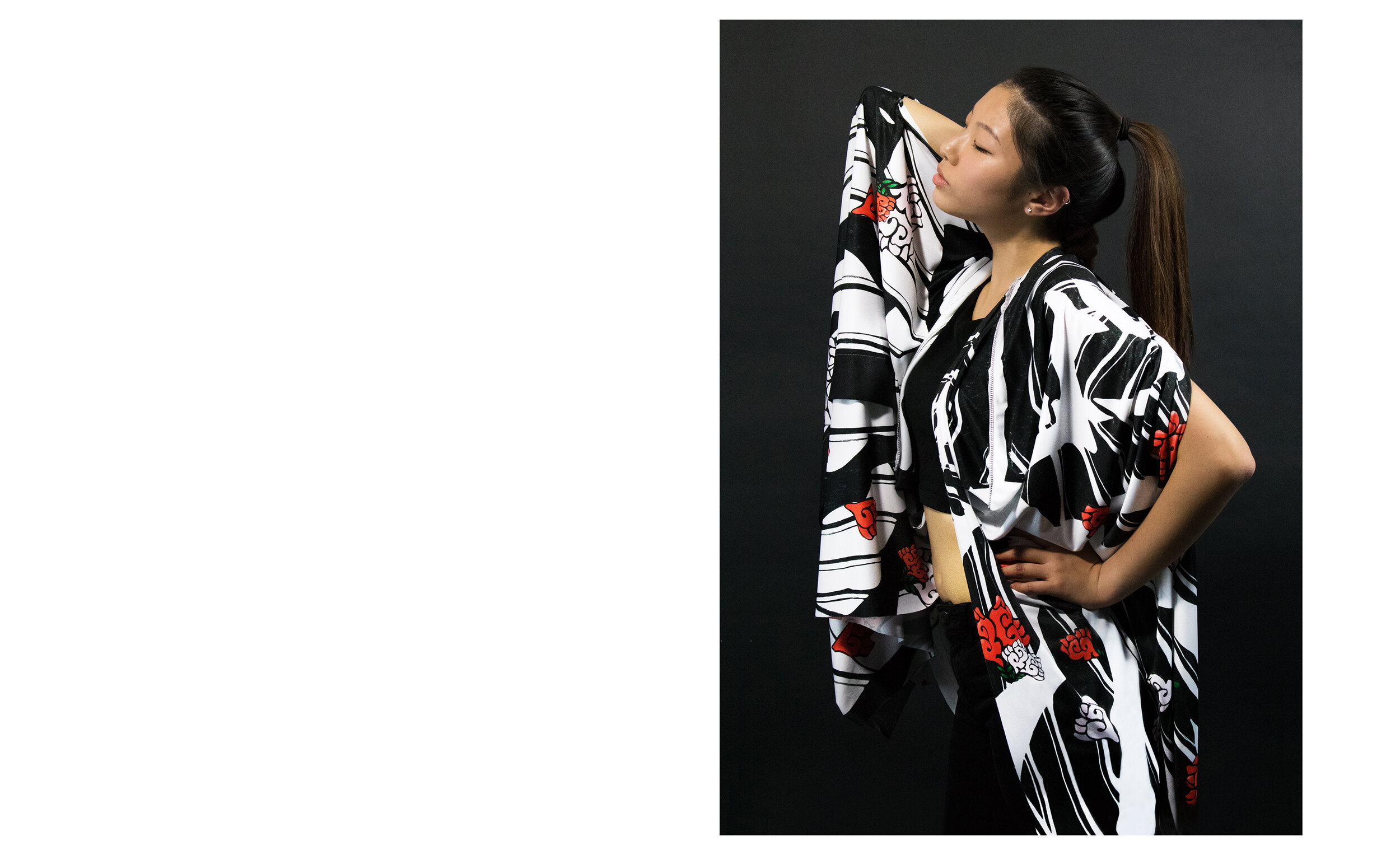 A woman with long black hair tied in a ponytail, wearing a black T-shirt and a black-and-white patterned robe with red floral designs, is standing against a black background with her eyes closed, one arm raised behind her head, and her other hand res