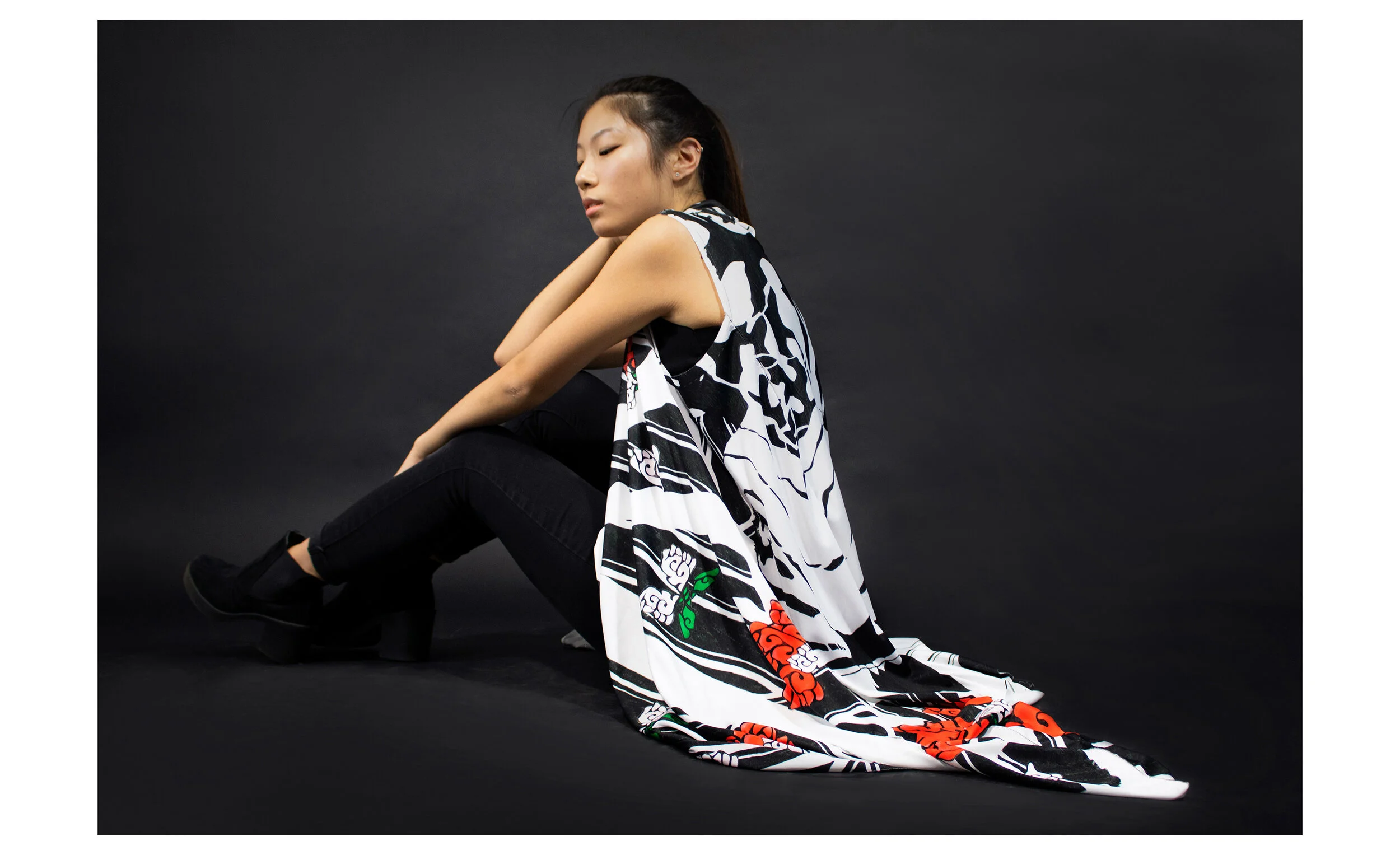 A woman with long black hair sitting against a black background. She is wearing a sleeveless top and black pants. Her eyes are closed, and she appears to be in a reflective pose.