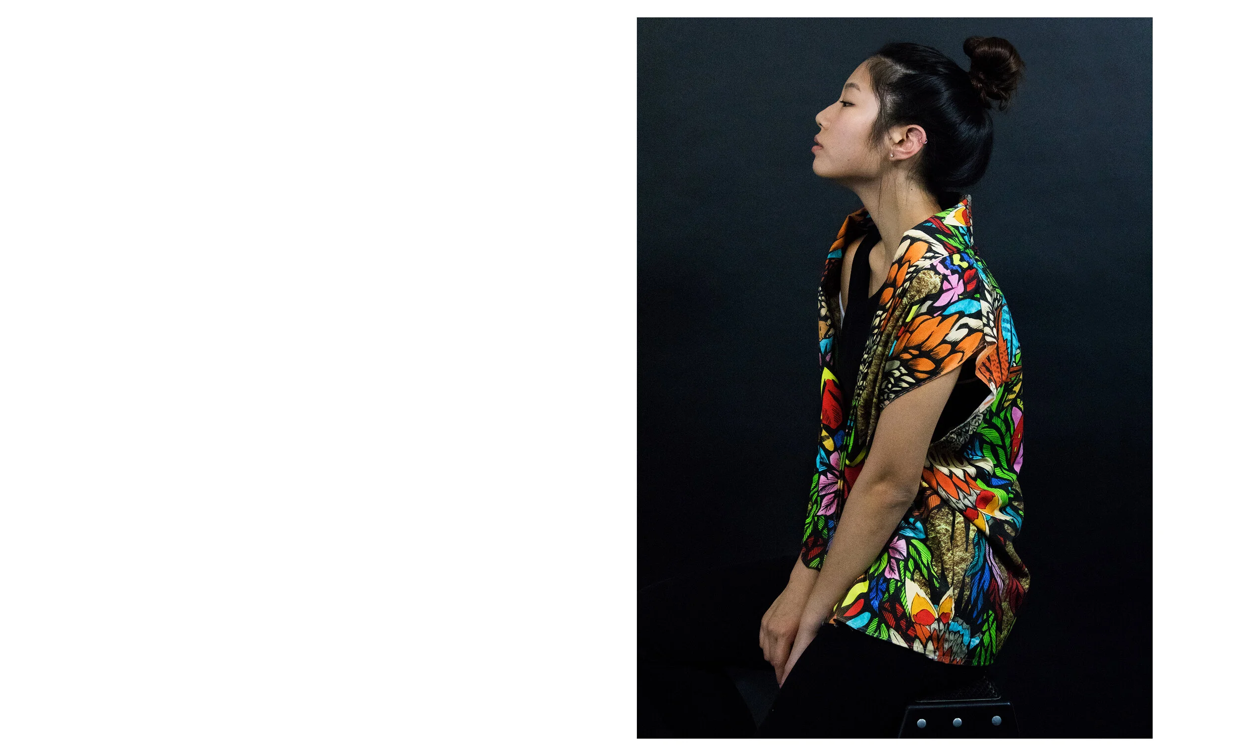 A woman with dark hair styled in a bun, wearing a colorful, patterned dress with floral motifs, is sitting against a dark background in a profile pose.