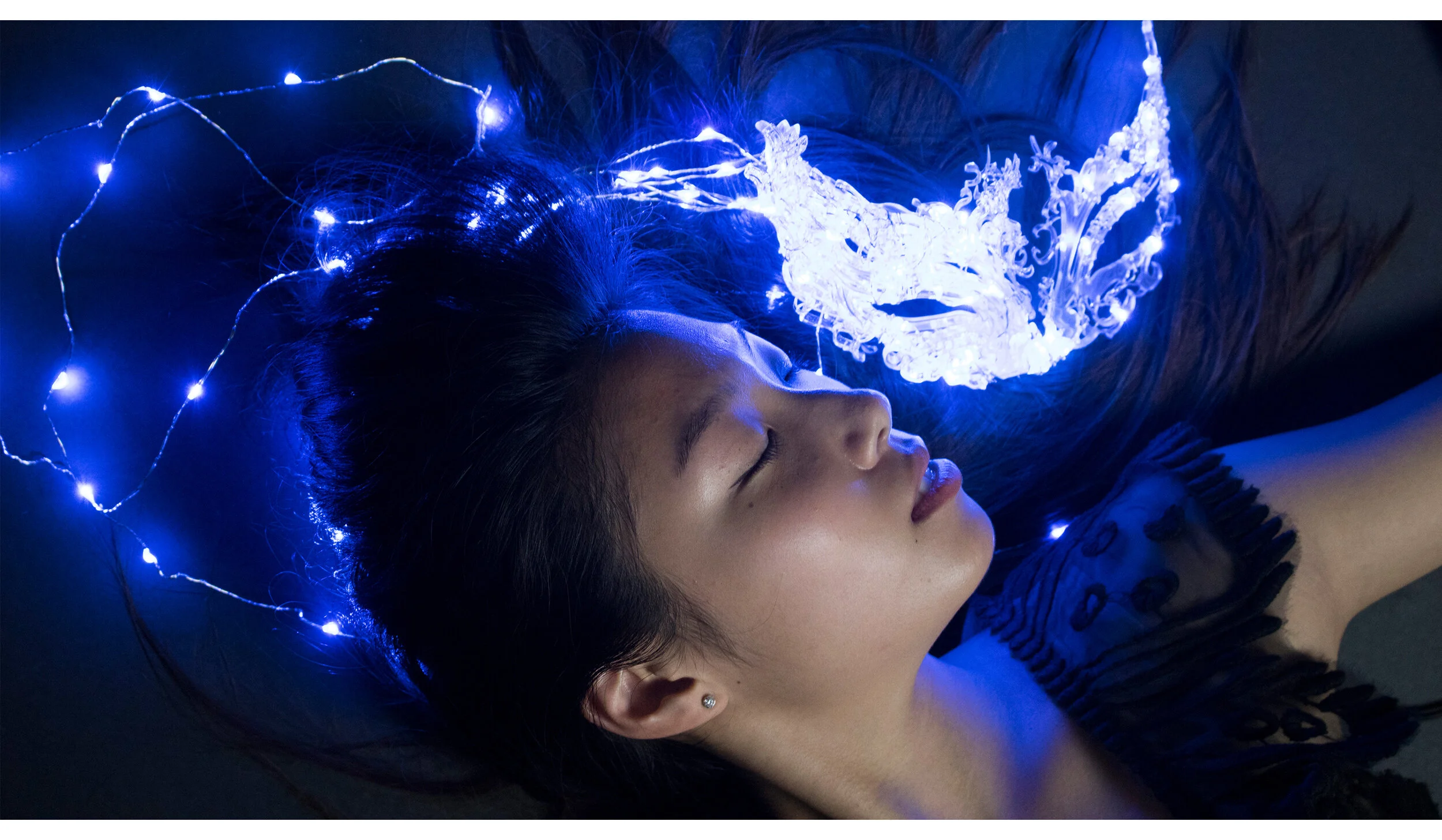 A woman with closed eyes lying down, surrounded by blue fairy lights and a glowing crystal decorative piece on her chest.