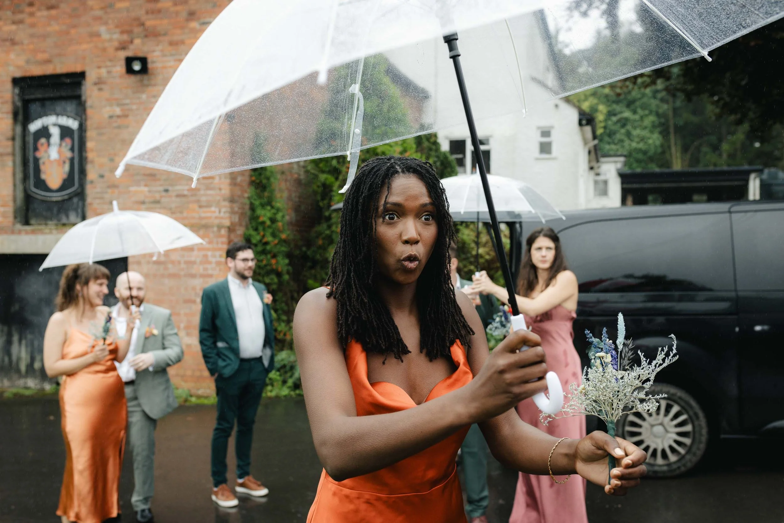 Bridesmaid sheltering under an umbrella, looking shocked at the rain as they arrive for the ceremony.