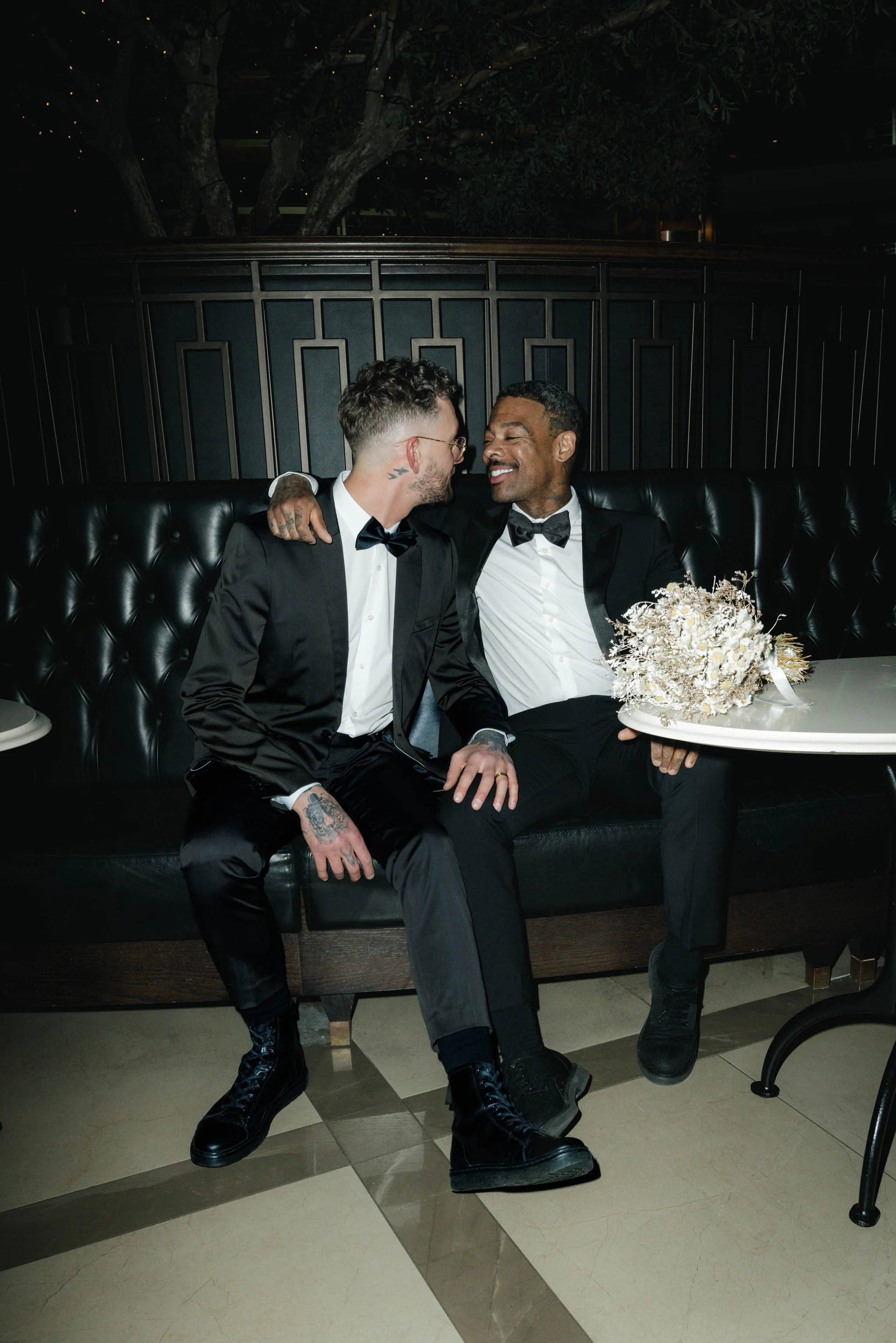 Two grooms in tuxedos sat at a city centre champagne bar.