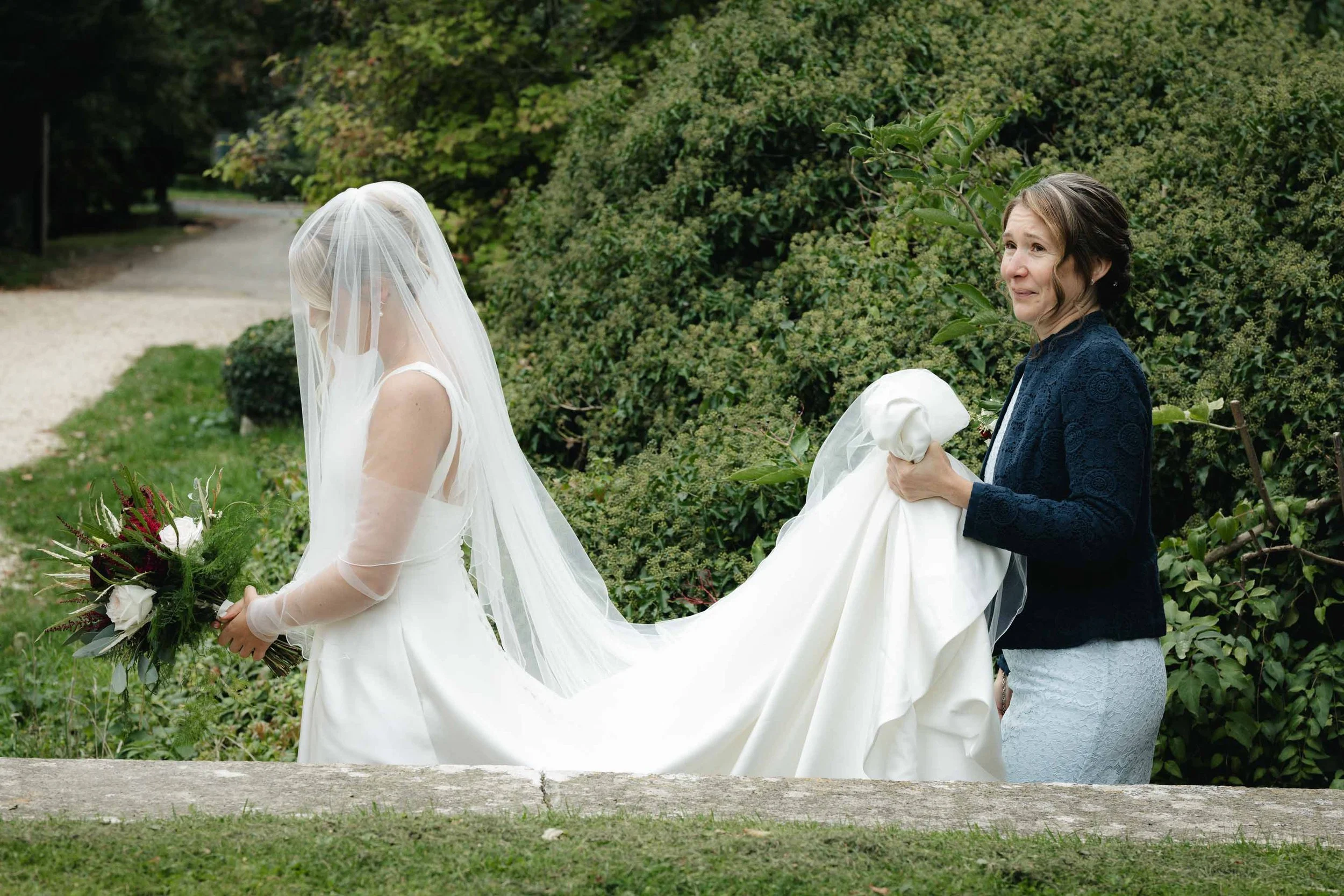  Documentary photo showing woman walking behind a bride, carrying her veil in her hands. 