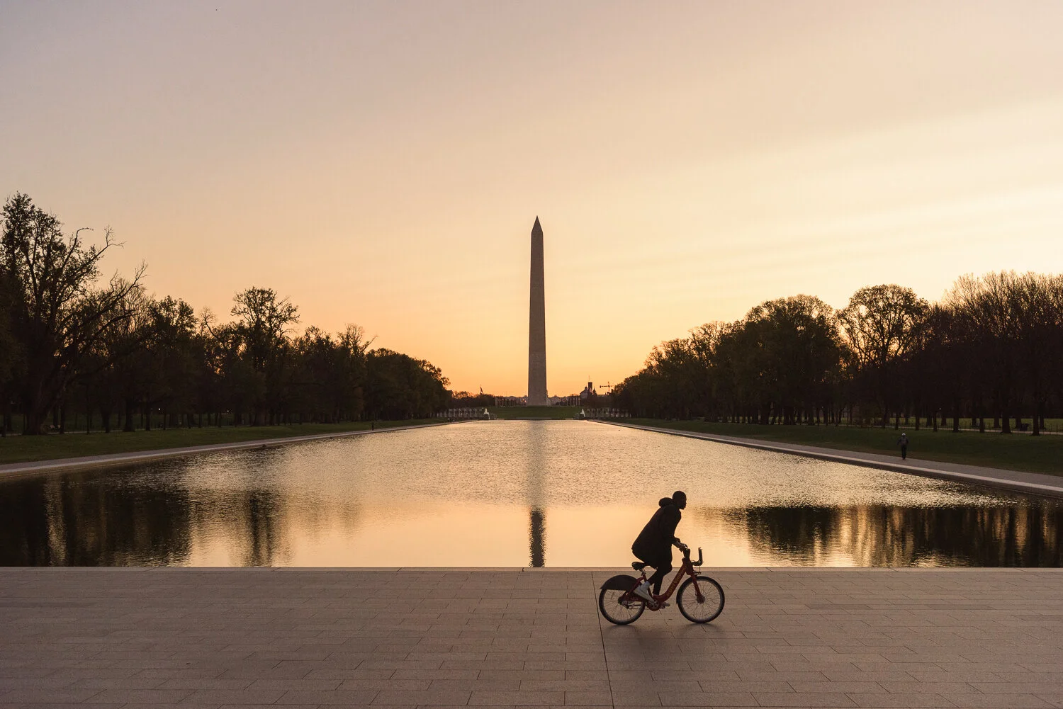 A man bikes in front of the Lincoln Memorial Reflecting Pool and Washington Monument at sunrise.