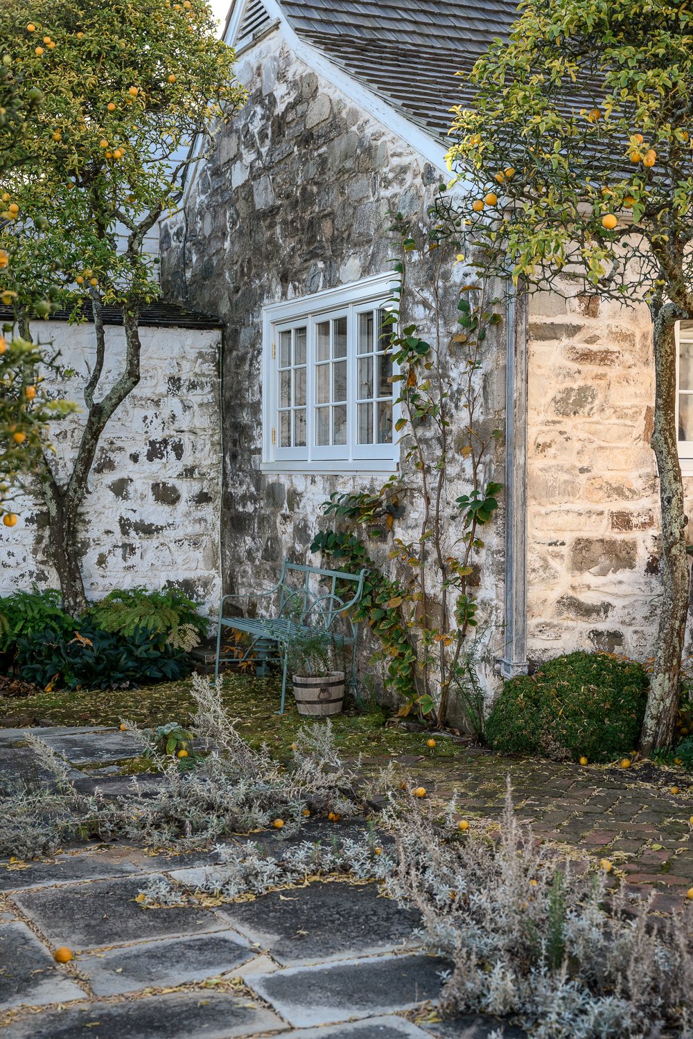 A rustic stone house exterior with a white-framed window, surrounded by greenery and lemon trees, a metal garden bench, and paved stone pathway with fallen lemon fruits.