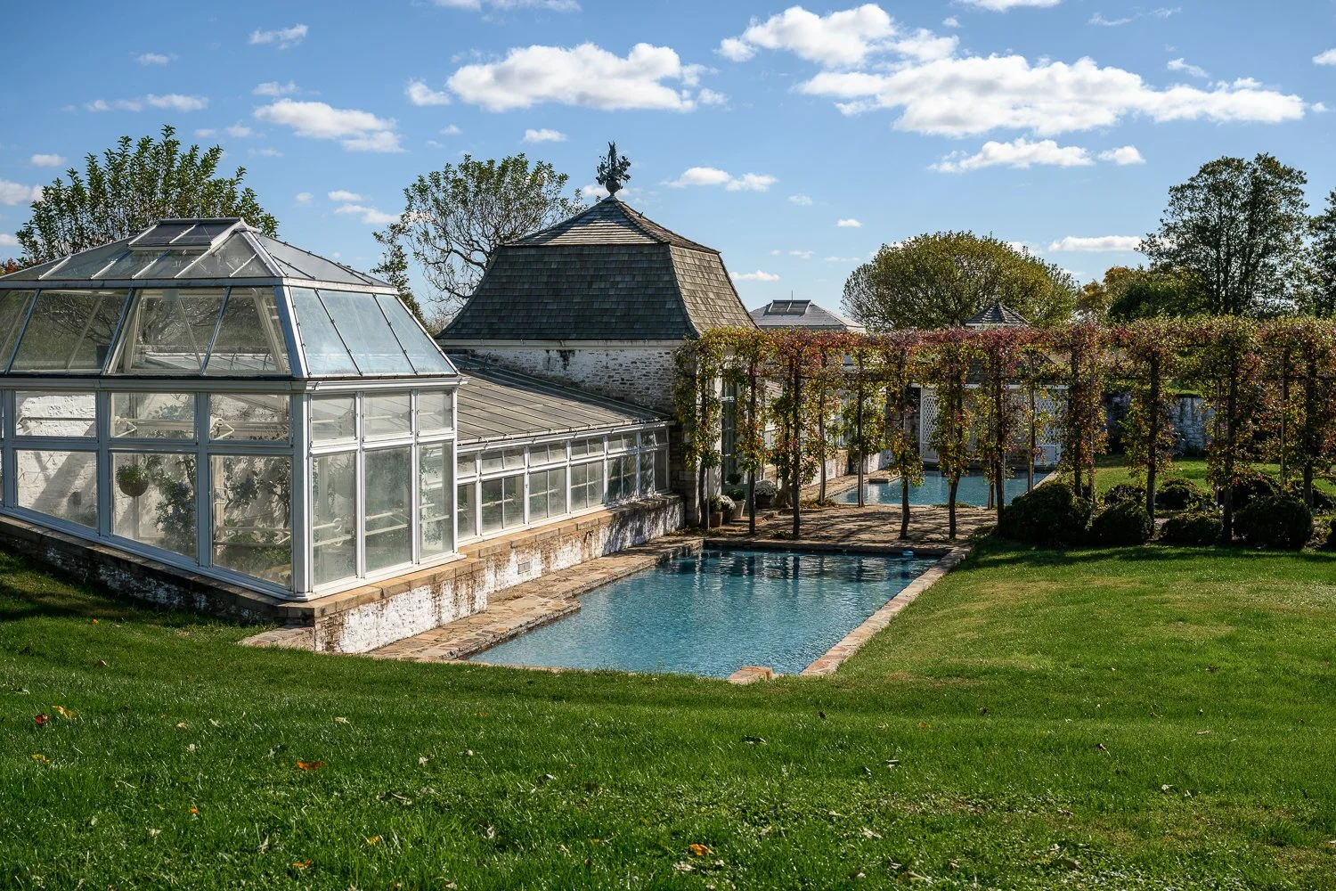 A garden scene with a glass greenhouse, a small swimming pool, a vine-covered pergola, and a stone building with a conical roof, under a partly cloudy sky.