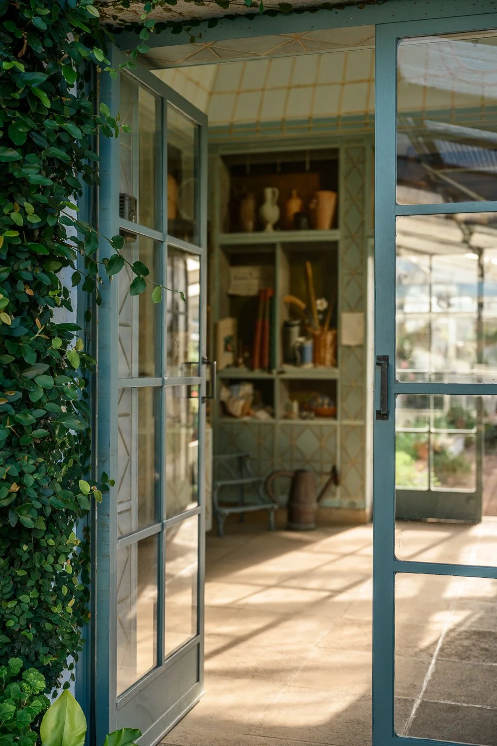 Open glass doors leading into a sunlit room with shelves containing pottery, books, and garden tools, with a lush green vine growing along the doorway.