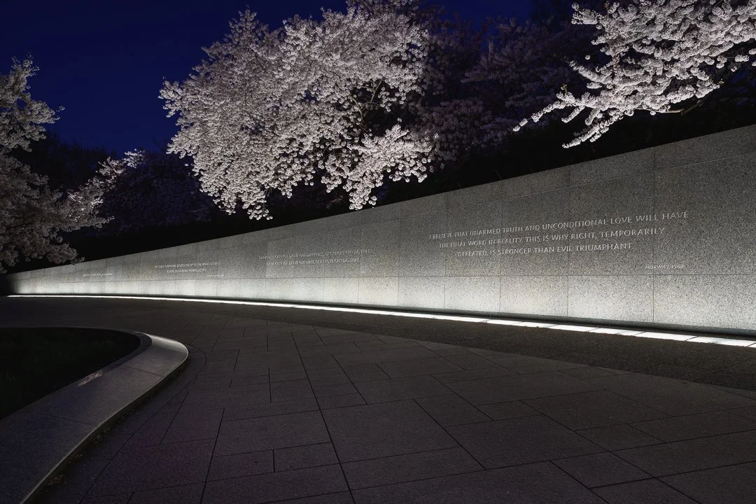 Nighttime view of a stone wall with inscriptions, illuminated from below, with blooming cherry blossom trees in the background. This monument is the Martin Luther King Jr Memorial in Washington, D.C.