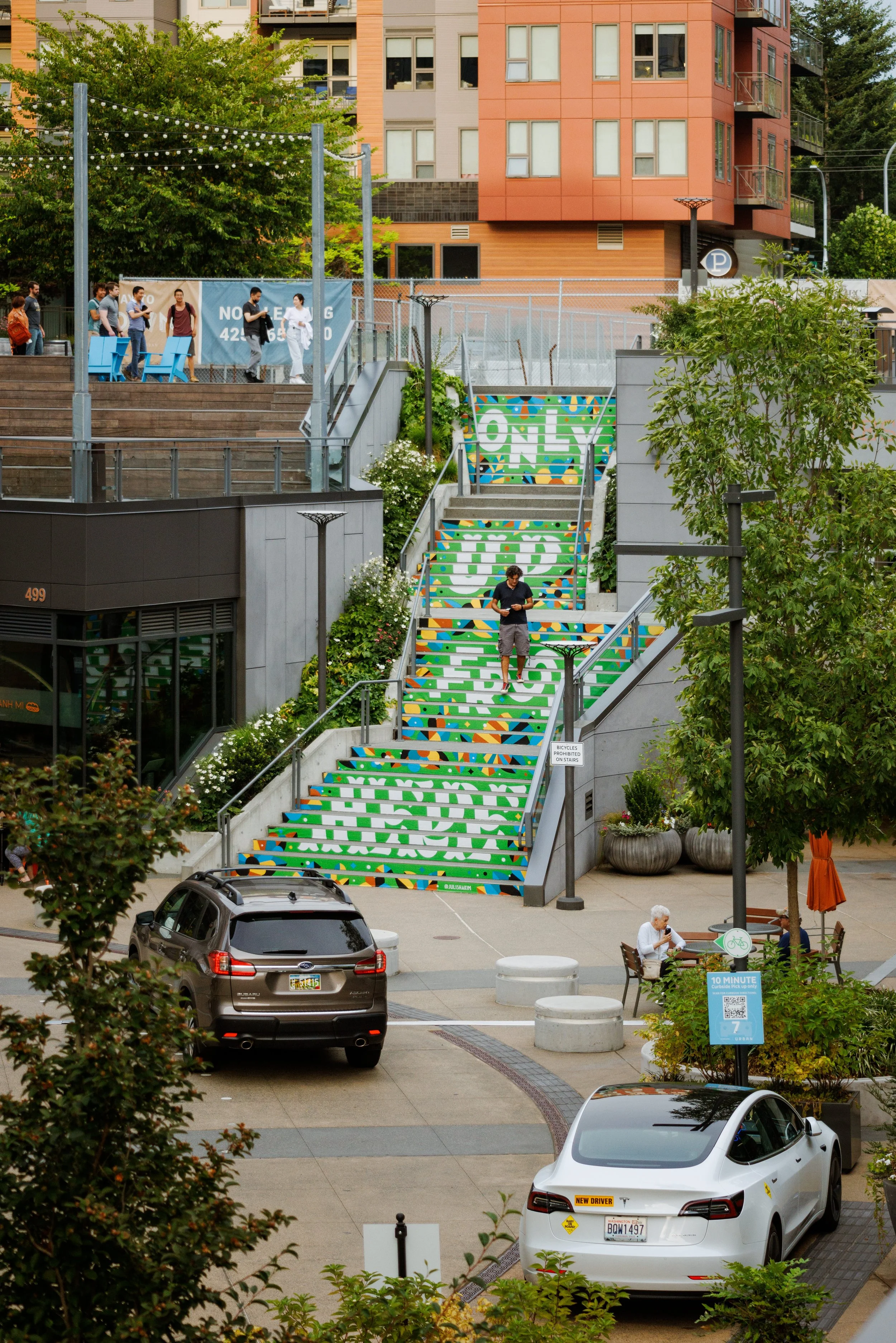 An image showing vinyl stair mural located in Kirkand Urban shopping complex. Mural design features hand lettered phrase of "Only Up From Here" with botanical illustrations around the phrase. There's a man walking down the stairs.