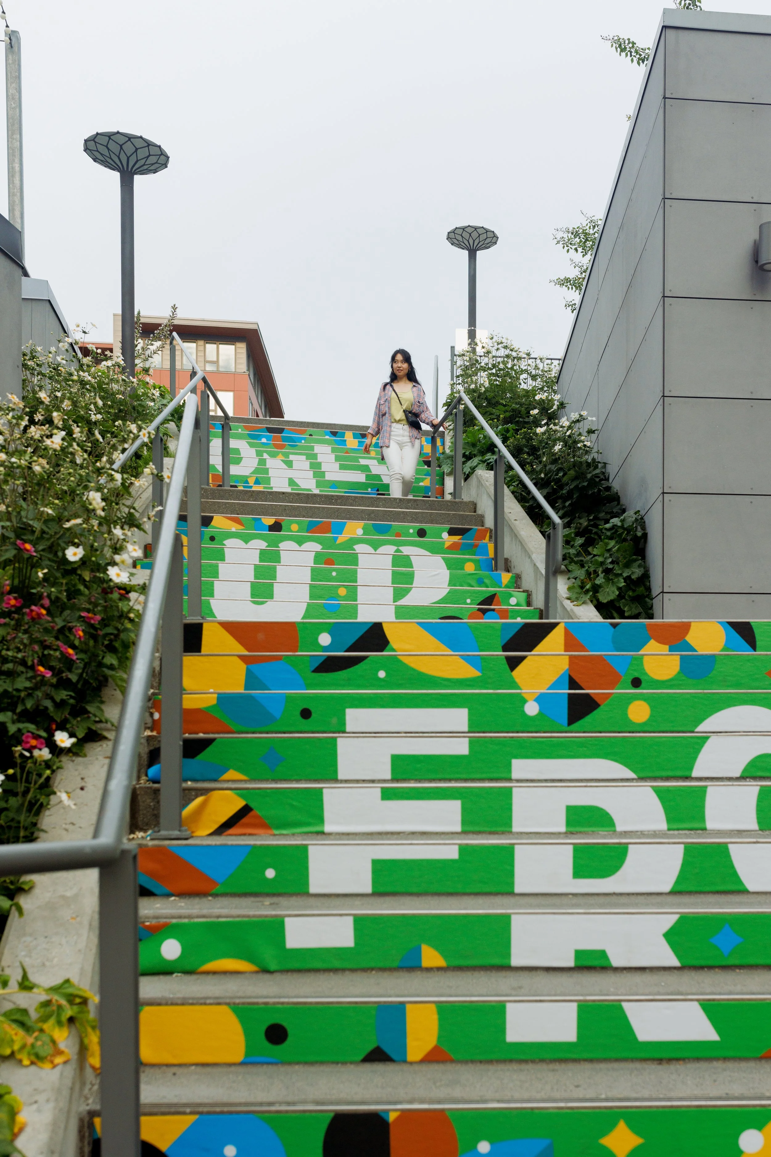 A close-up image showing vinyl stair mural located in Kirkand Urban shopping complex. Mural design features hand lettered phrase of "Only Up From Here" with botanical illustrations around the phrase. There's a young Asian woman walking the stairs.