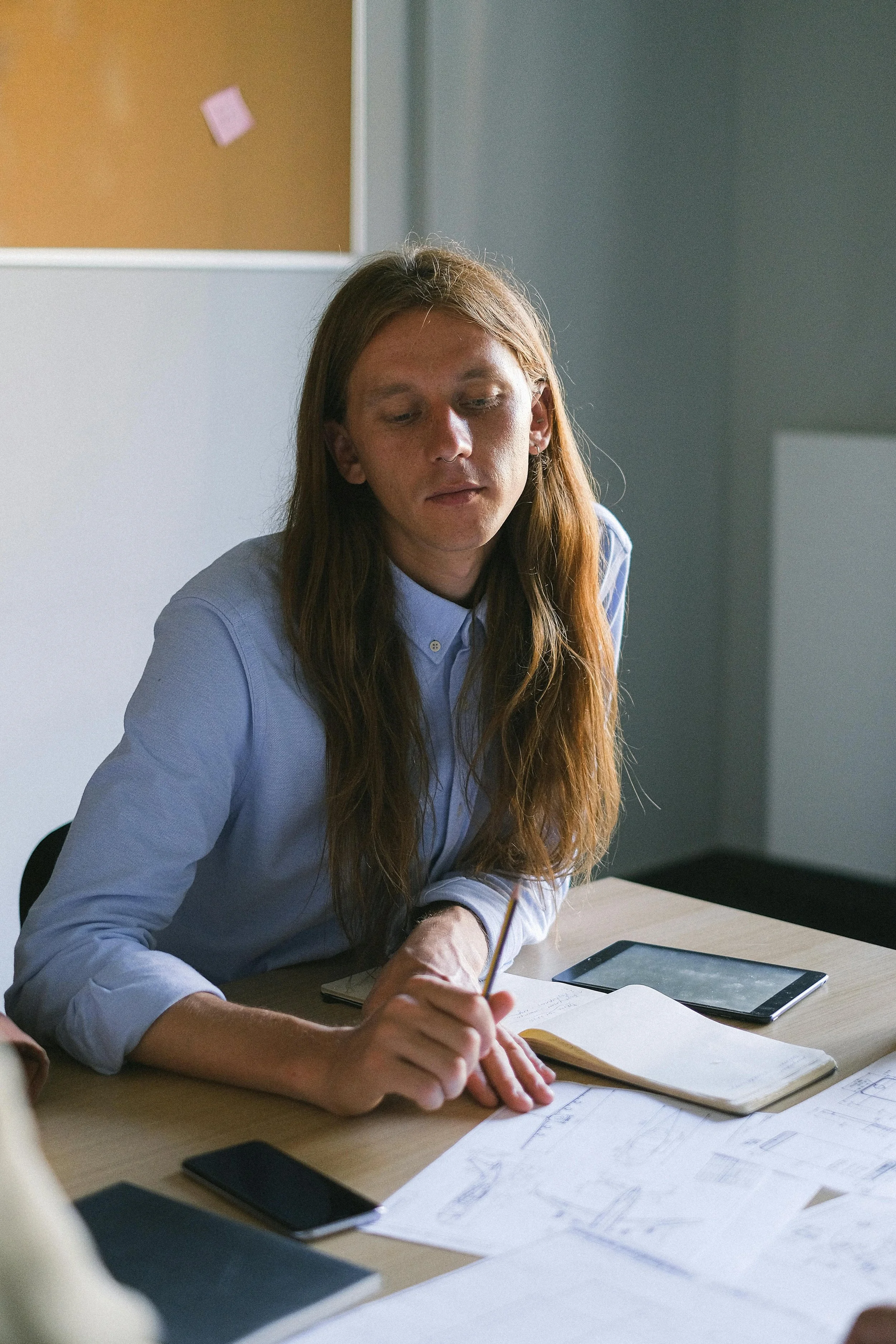 A woman with long red hair wearing a light blue shirt, sitting at a desk with papers, a tablet, and two smartphones. She is holding a pencil and appears to be reviewing documents or sketches.