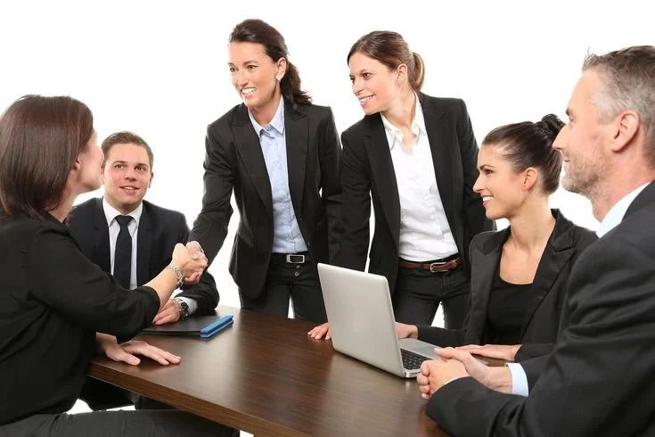 Five professionals in business attire engaged in a meeting, with two women standing and three others seated, one shaking hands and one using a laptop.