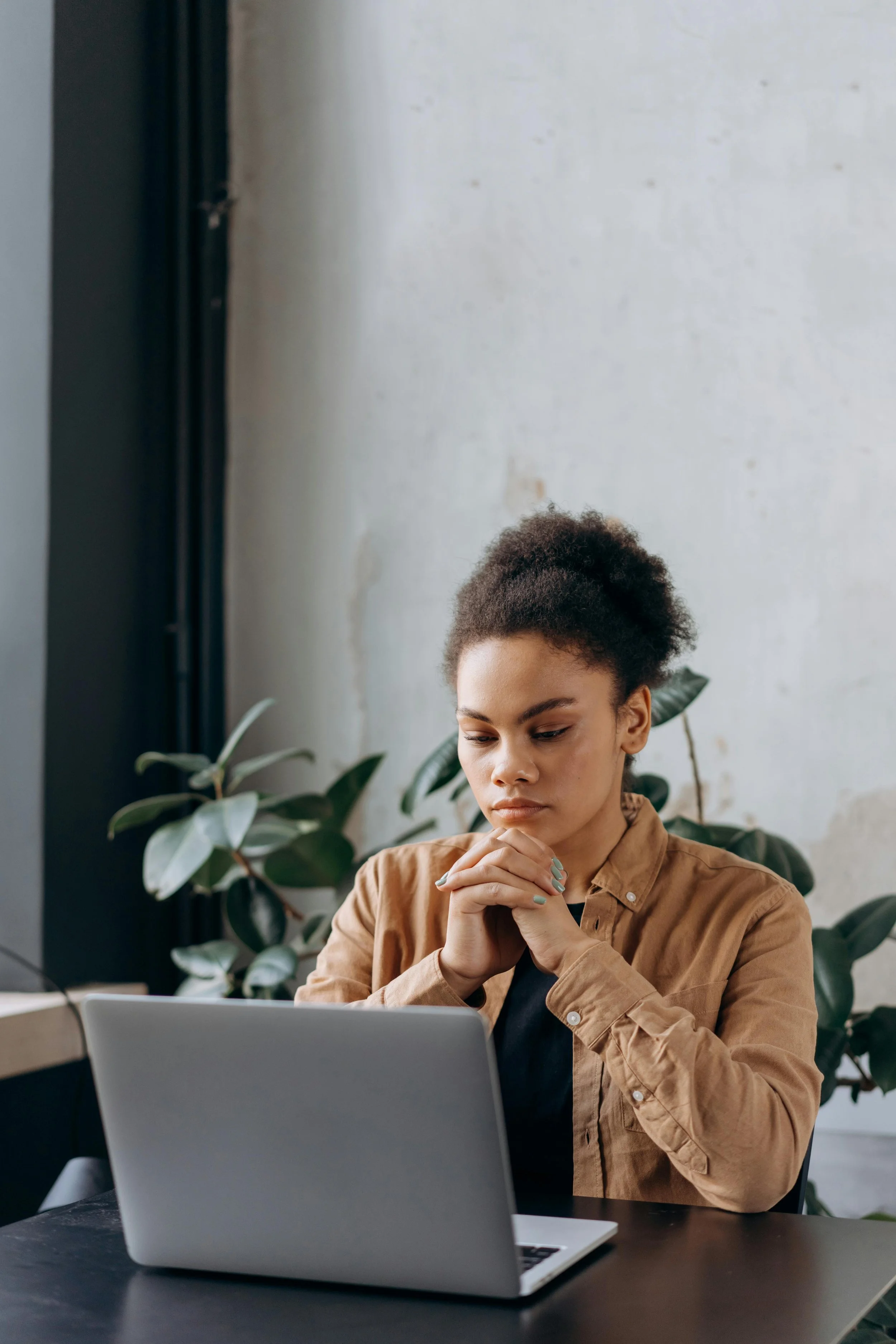 A woman sitting at a desk with a laptop in front of her, deep in thought, with green plants and a white wall in the background.