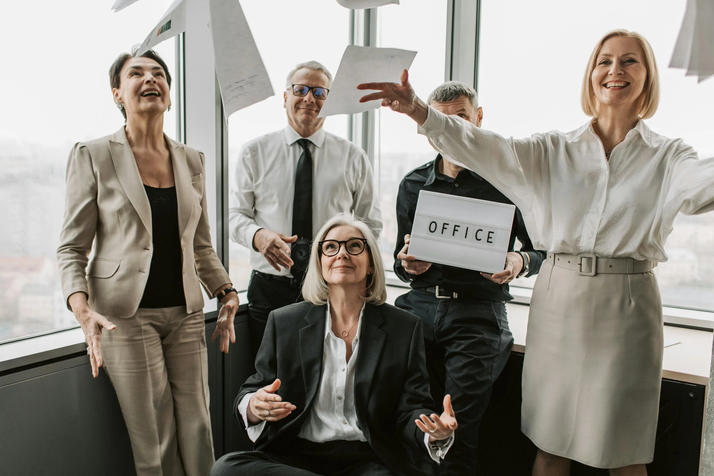 Group of five diverse business professionals in an office, some standing and one woman seated, with papers and a sign that says 'OFFICE,' engaging in lively conversation and laughter near large windows.