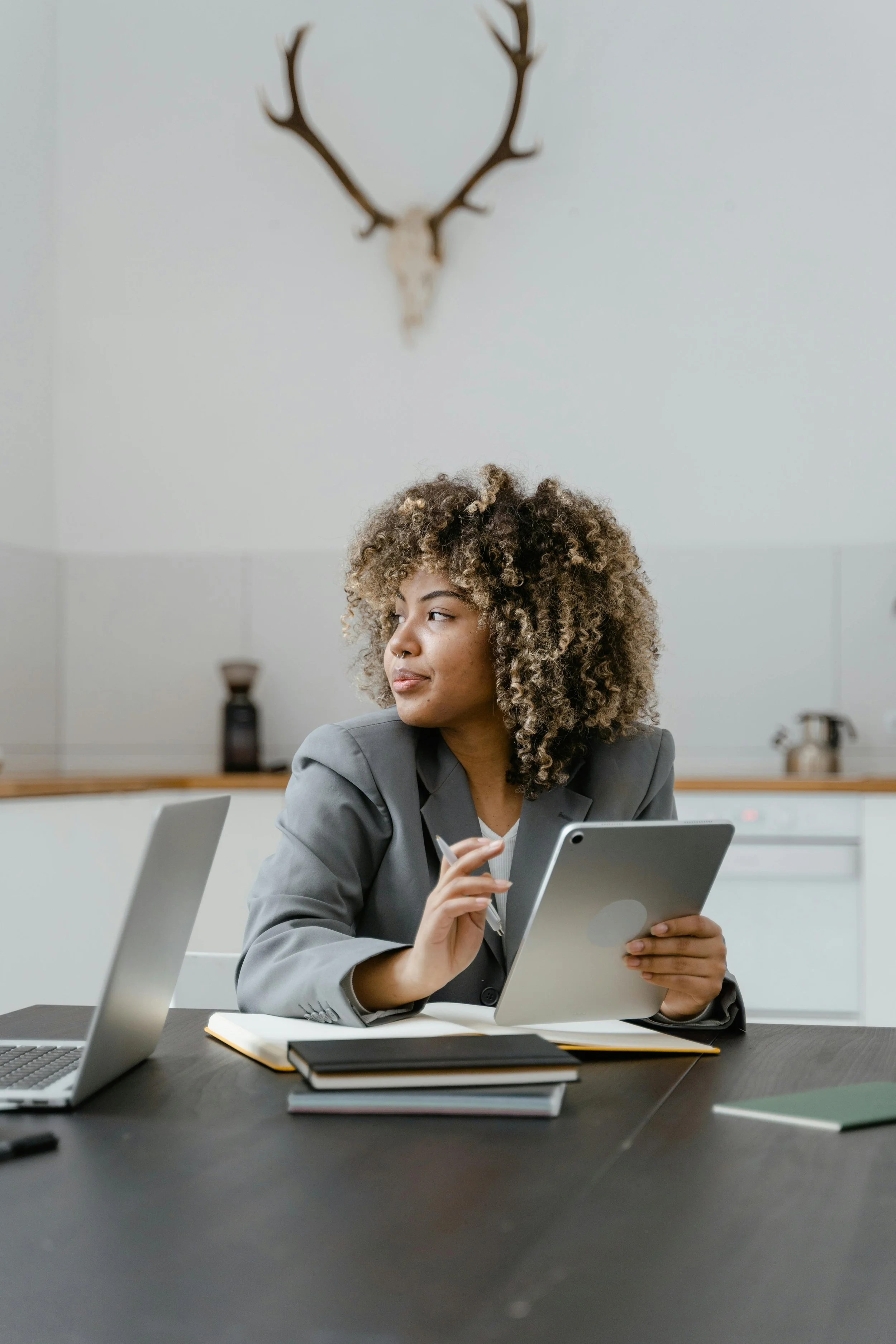 A young woman with curly hair is sitting at a desk with a laptop, notebooks, and a tablet, looking to her left in a modern kitchen with a deer skull with antlers mounted on the wall behind her.