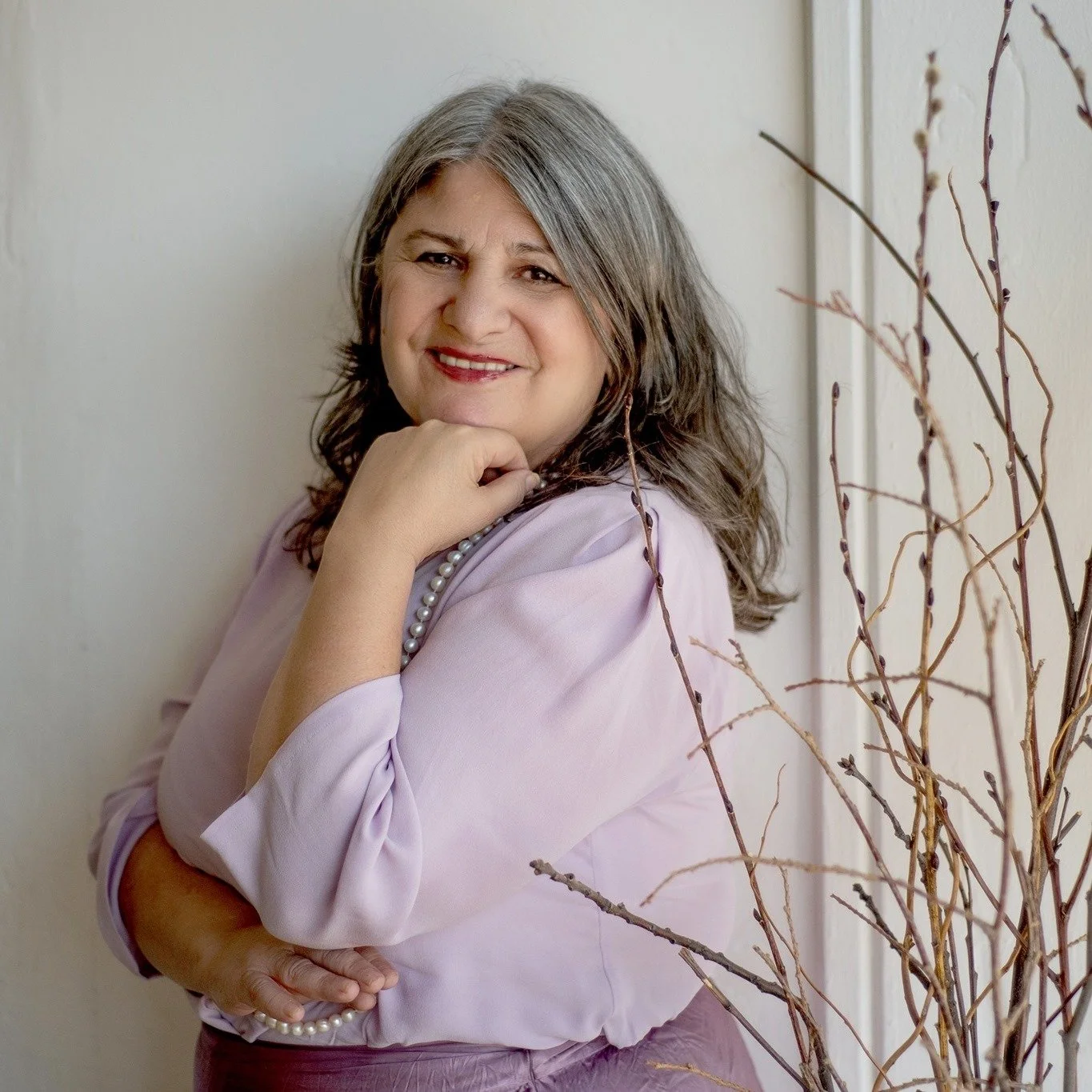 A middle-aged woman with shoulder-length gray hair, wearing a light pink blouse, a pearl necklace, and smiling while looking at the camera, standing next to a light-colored wall.