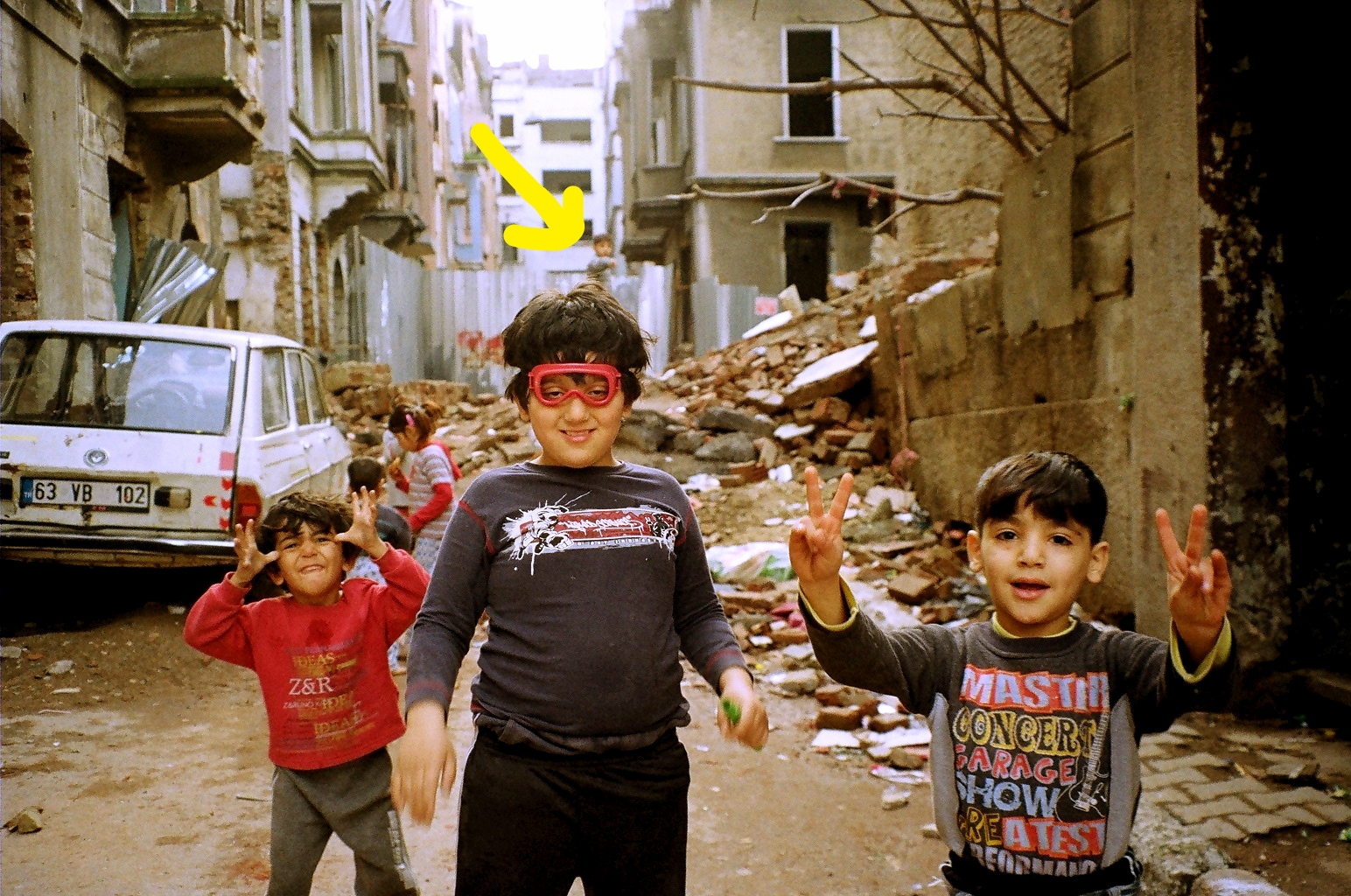 Tarlabaşı neighbourhood of Istanbul, Turkey. Photograph Sila Yalazan, 2013. Author: S.T. Lore Photographer: Sila Yalazan Client: ELP Journal Designer: Rob Milne Format: Postcard, 100 copies Year: 2015 STORM GODS OF SYRIA Three Syrian Kurdish refugees pose beside a Renault-12 TSW Station Wagon. One child mocks the viewer: his teeth are bared, his hands are in the shape of horns. Another stands forward and offers peace: his arms are held high and open wide. The largest (wearing goggles) stands central: he is benevolent and sturdy and looks prepared for rain. The same model Renault-12 featured in the opening scenes of the 2012 James Bond movie SKYFALL . A motorbike chase raced through Istanbul toward the city of Adana before reaching a tense final shoot-out on the Varda Viaduct. One of 22 train tunnels breaching the Taurus Mountains, they were dug out by Germany in 1912 as part of the Istanbul-Baghdad railway. Crossing the natural territory of the Kurdish people --- steep mountain ranges separating four warring nations --- previously the journey required a treacherous pass through the Cilician Gates using bullock drawn caravans, confronting violent thieves, rock collapses, and death. Now, Renaults and Jeeps merge onto the Class-A European highway called the E-90 which plummets tourists from Istanbul to Cappadocia. The trains are filled with pensioners and the nostalgic. Refugees traverse the territory anyway they can. In a Turkish only re-branding, the Renault-12 was marketed as the Renault ' Toros' . Named after the symbolic bull of the Taurus Mountains in reference to the mythical transport of the Babylonian storm god Hadad . Also known as Baal , the ancient god of rain and of oracles is depicted on religious cylinder seals 'standing on the back of a bull, brandishing a mace or another weapon in his right hand and a thunderbolt in the other…he is bearded, wears a horned head-dress, and treads the mountain tops at the feet of waves '. Son of the great sky god Anu his temples of worship are secured to the mountaintops and decorated in thunderbolts and bulls. Appearing as a demon in the Marvel Comics , this cartoon image, like all myths, explains the forces beyond our control. Turkish Radio and Television Corporation (TRT) started its 24-hour Kurdish television station with the congenial motto: ' We live under the same sky '. It is a poetic statement, full of inclusive sentiment, yet Kurdish cartoons were banned in Turkey until as recently as 2013, alongside the letters X, Q and W. This draconian political move, designed to curb Kurdish literacy, forced children into the rubble-strewn streets. No longer watching television, they played amongst the rocks and discovered a battered 'T oros '. Permanently sealed inside a photograph, these sky-fallen casualties instinctively mimic the ancient storm gods, yet one child alone remains defiant in the background. Standing atop a pile of rubble, he avoids the playfulness of the others and points directly at the viewer. His action, more menacing than a pose, could be curiosity, yet it could also be hostile: an omen, a premonition, a warning of another storm. 