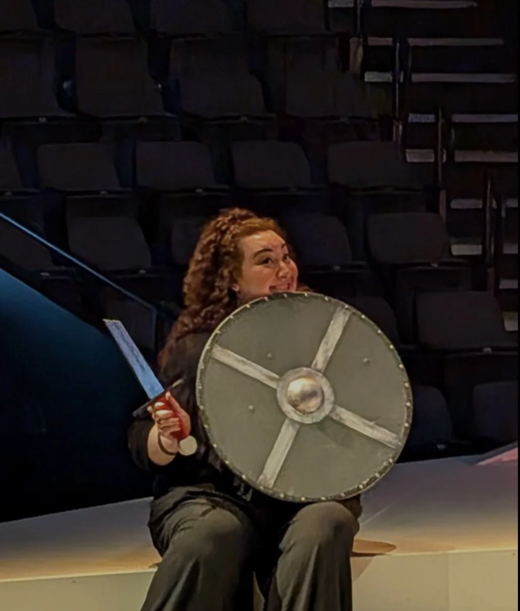 Jaclyn Grossman sits onstage holding a sword and round shield, smiling playfully during a rehearsal moment in an otherwise empty theatre.
