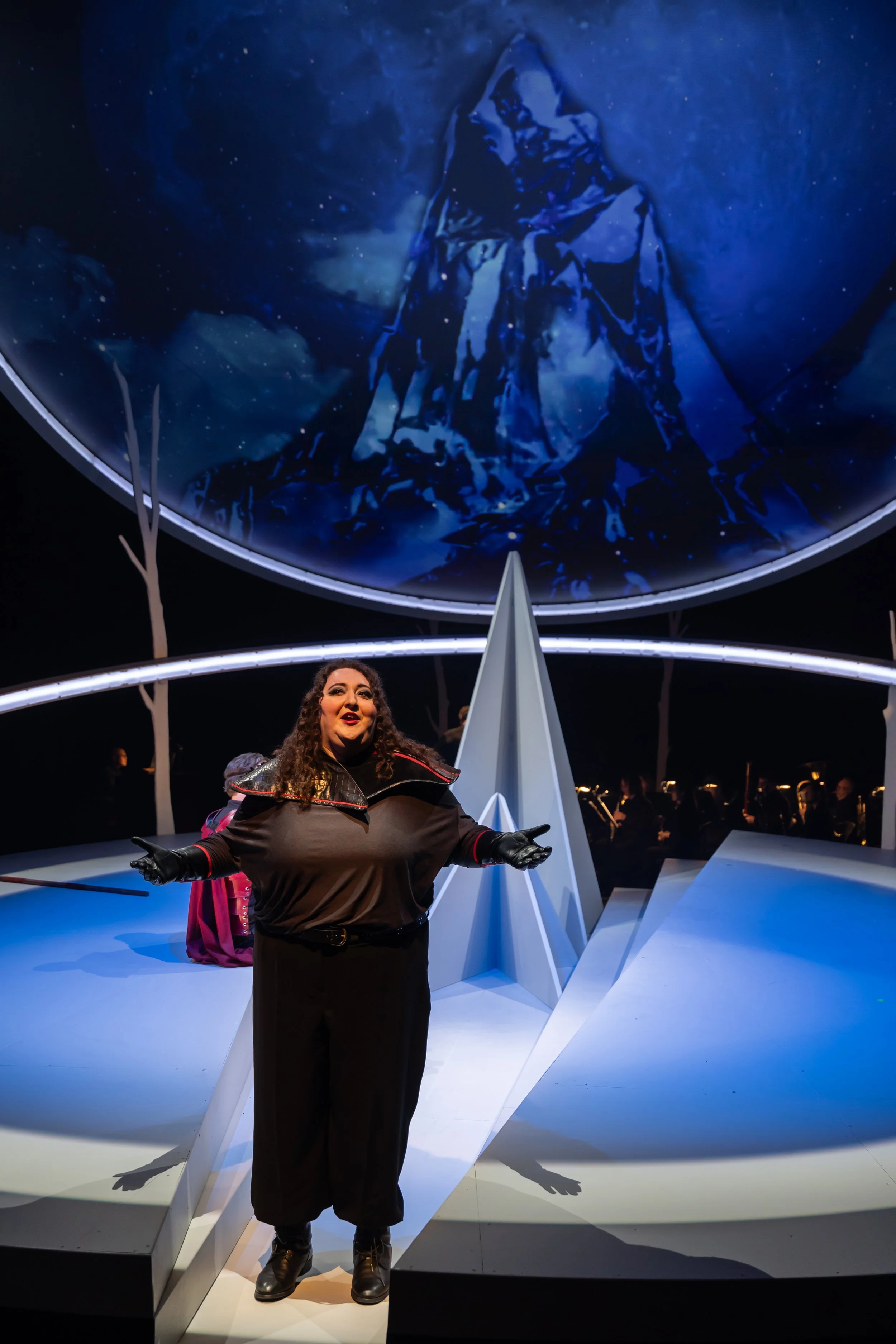Jaclyn Grossman as Brünnhilde stands centre stage with arms open, wearing a dark costume with structured shoulders, beneath a large circular projection above the set.