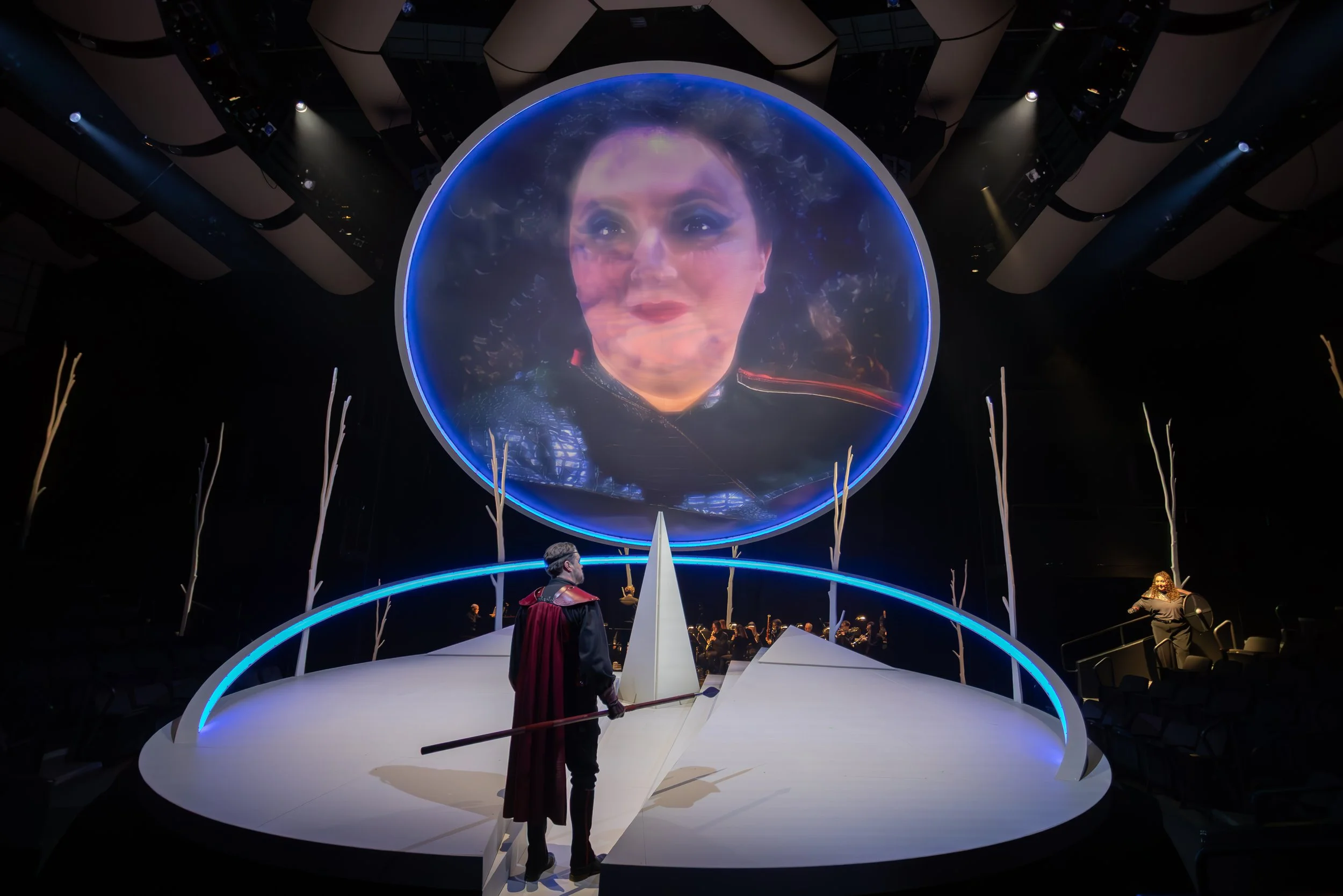 A large circular screen displays a close-up of Jaclyn Grossman’s face in character, looming above the stage while another performer stands below holding a spear.