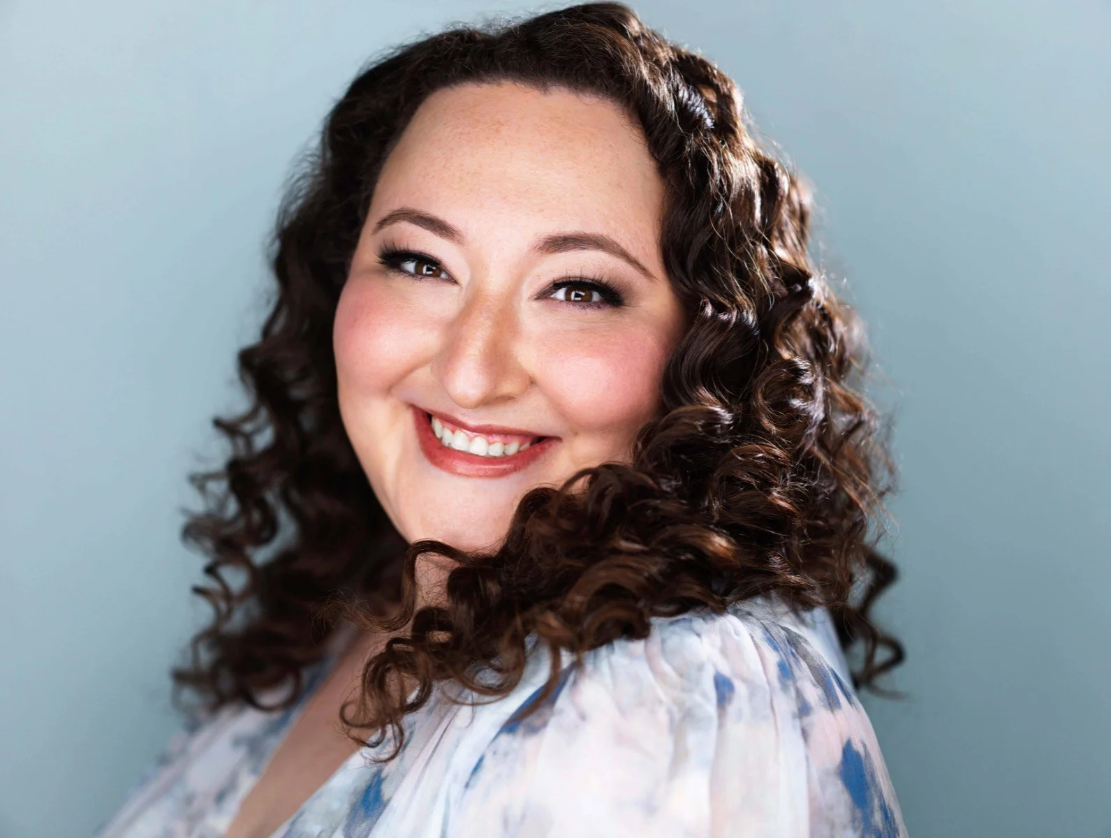 Headshot of soprano Jaclyn Grossman smiling toward the camera, with long dark curly hair and a soft blue-grey background.