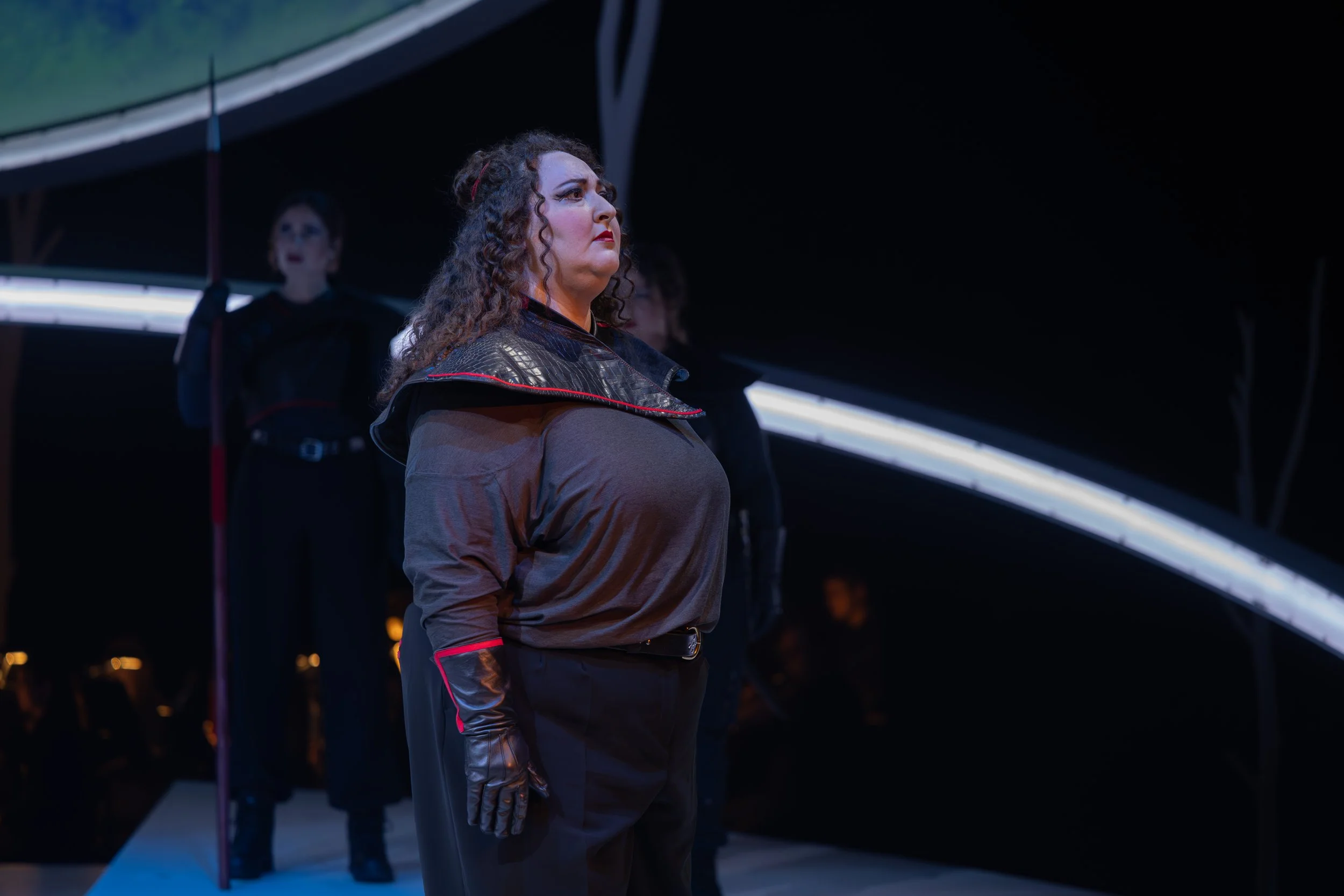 Jaclyn Grossman as Brünnhilde stands in profile onstage, wearing a dark costume with gloves, lit dramatically against a minimalist set.