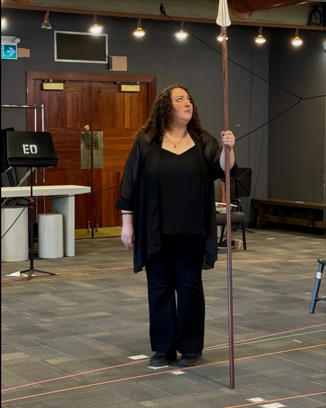 Jaclyn Grossman stands in a rehearsal space holding a long spear, looking upward as she prepares for a scene, with taped stage markings on the floor.