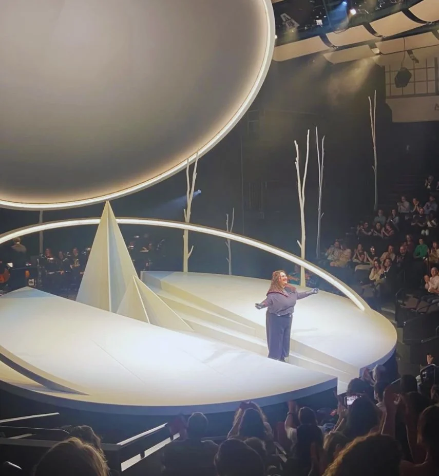 Jaclyn Grossman takes a bow onstage as Brünnhilde, standing on a minimalist circular set with a large glowing disc overhead, while the audience applauds.