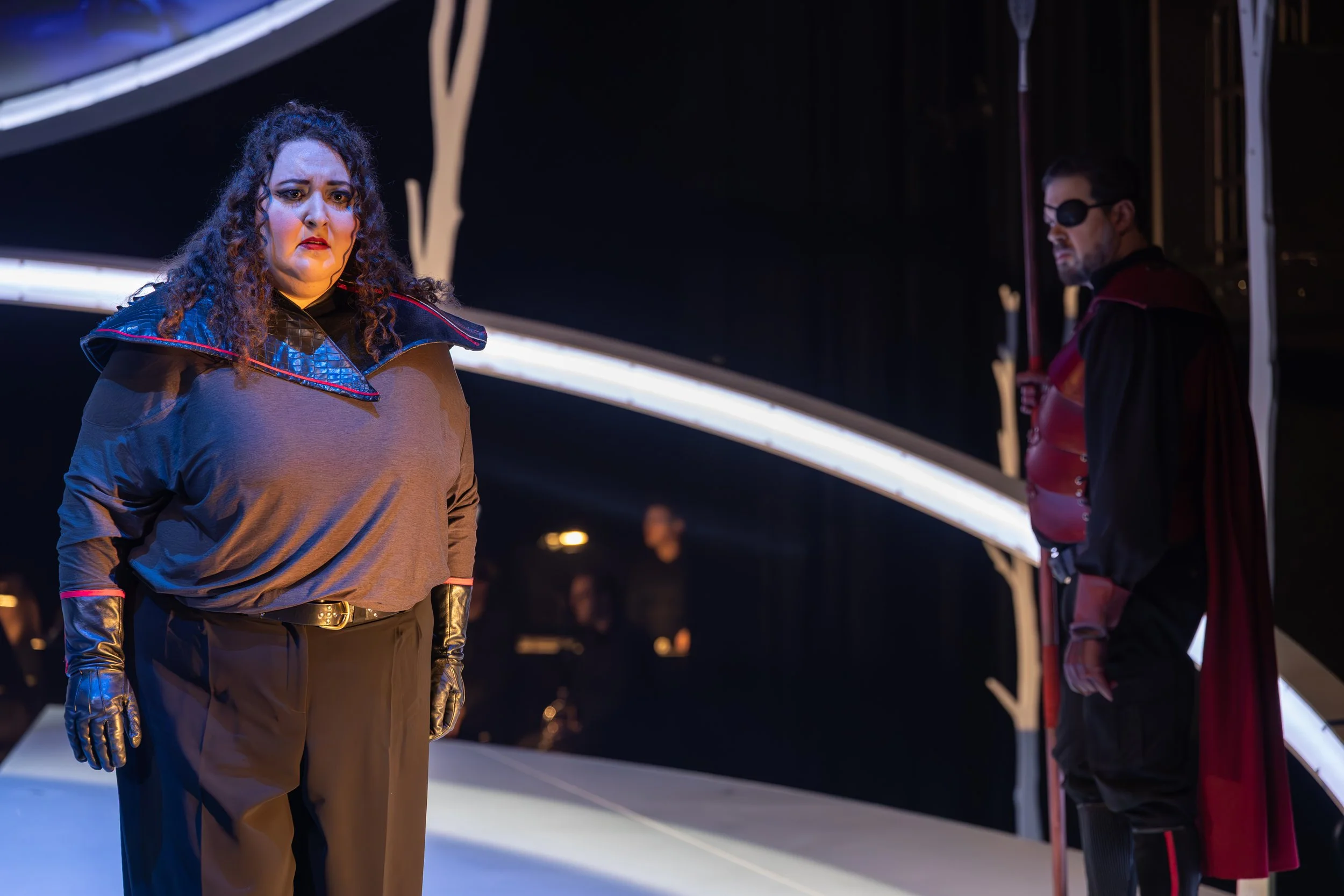 Jaclyn Grossman as Brünnhilde stands in the foreground looking distressed, while another performer dressed in red and black stands behind her holding a spear.