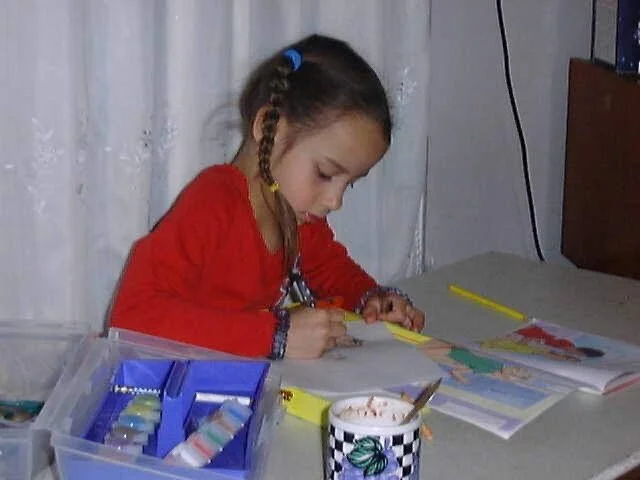 A young girl with braided hair, wearing a red shirt, sitting at a desk, coloring in a book with crayons.