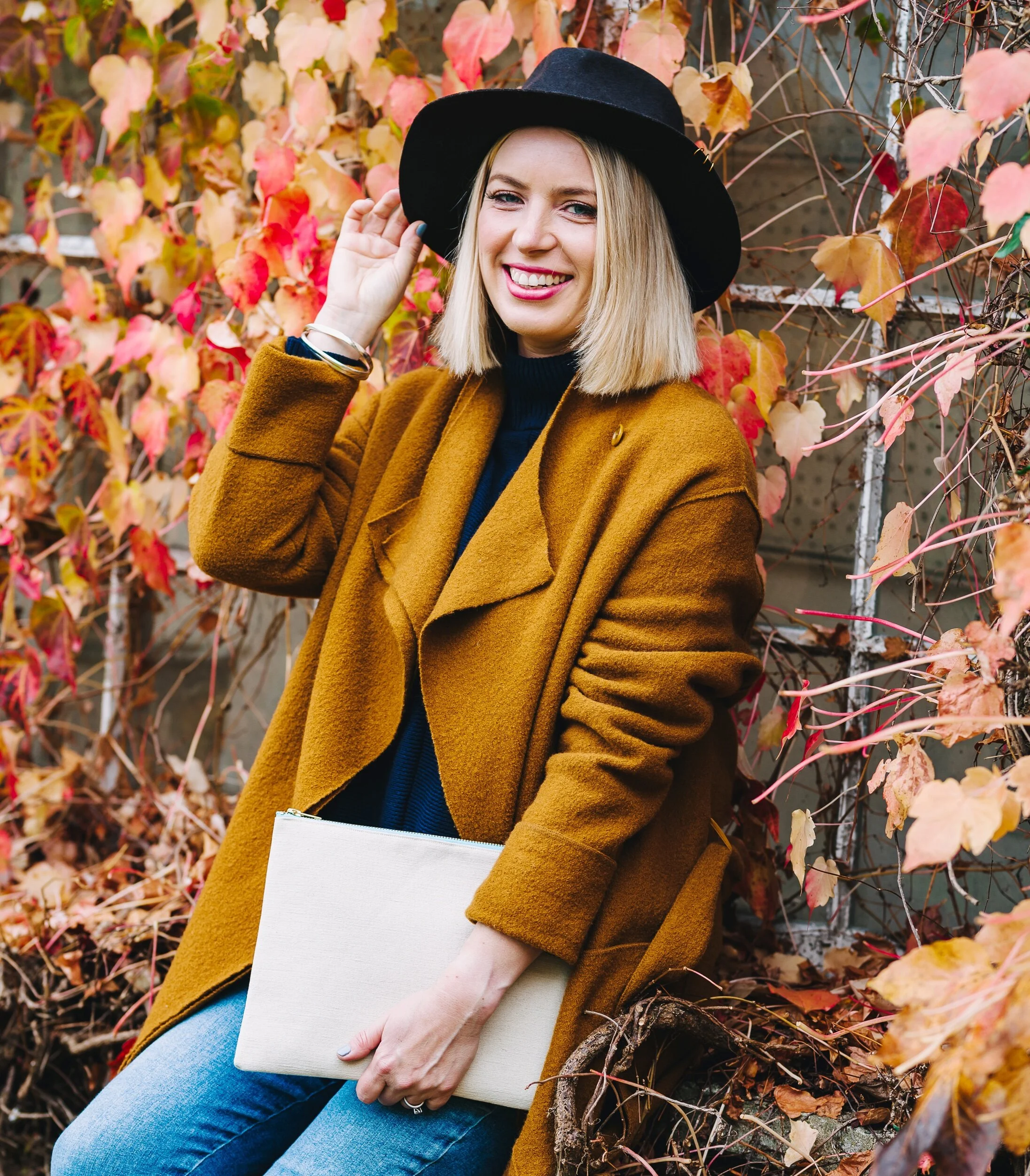 Elizabeth wears a sustainably friendly wool coat and  hat perching with autumnal leaves behind he and holding a B Collection large clutch bag she is smiling Carry Your Cause
