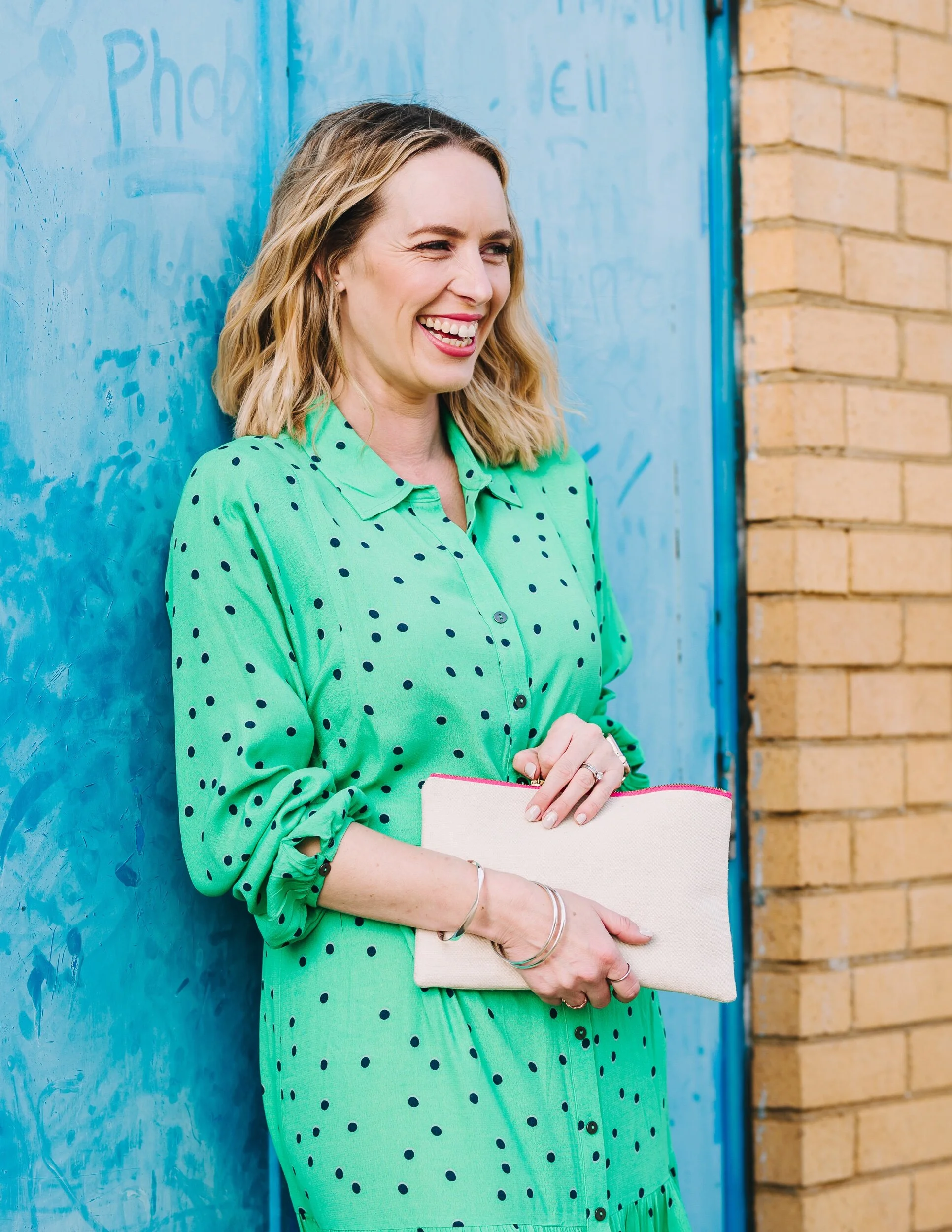 Elizabeth wearing green spot dress stands in front of blue door brick wall smiling holding cream hemp clutch bag bright pink zip