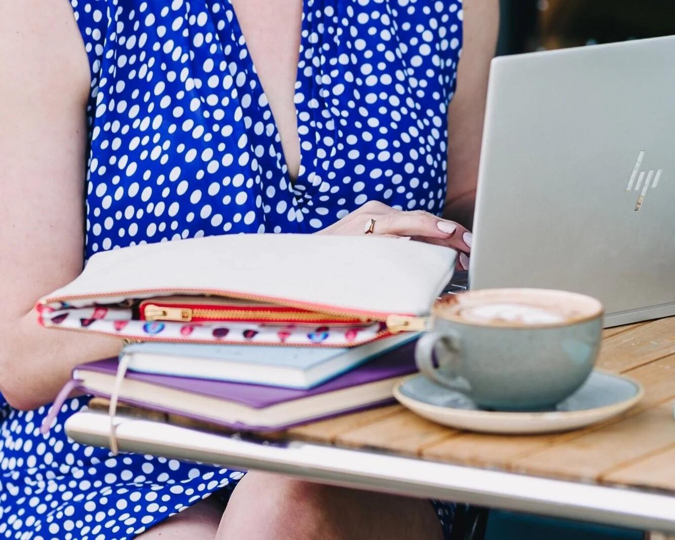 woman wearing blue and white dress sat at cafe table with coffee cup laptop V Collection clutch bag laying on notebook