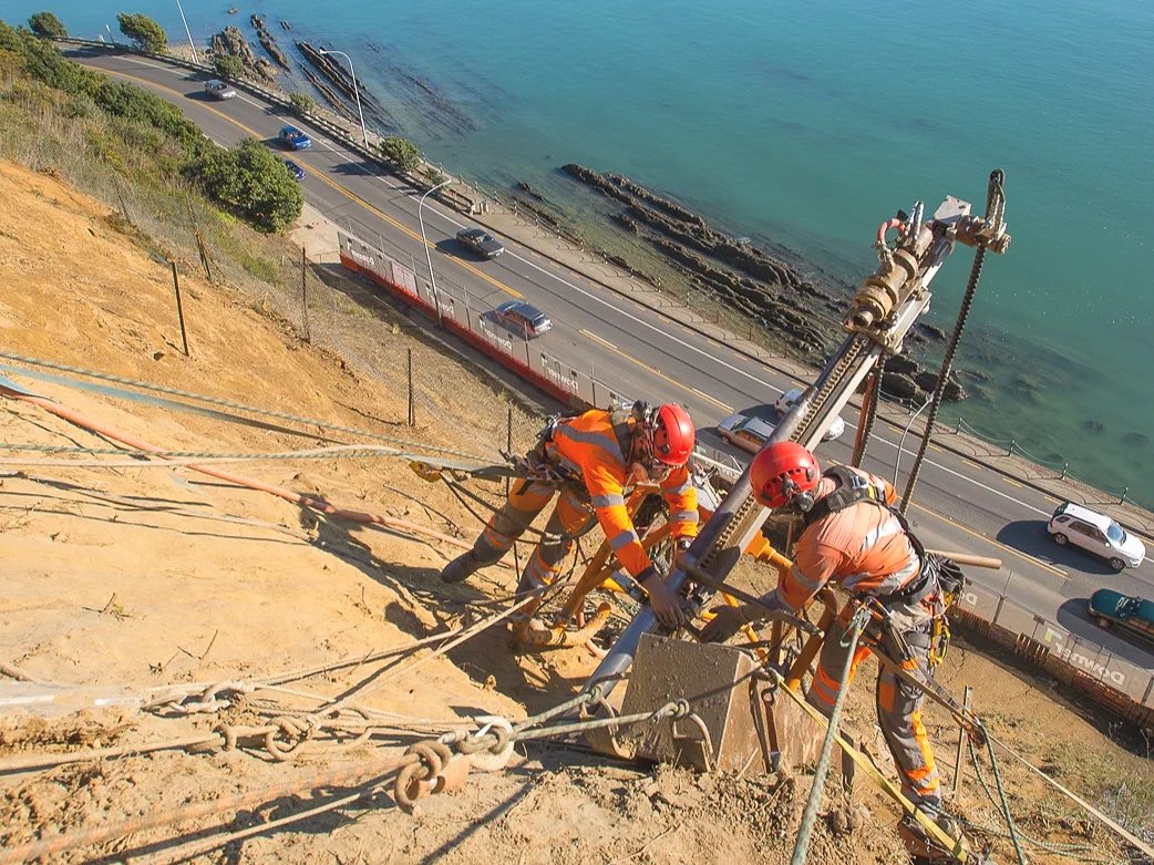 Two workers wearing safety gear, including helmets and harnesses, are securing equipment on a steep hillside above a coastal highway with cars.  Nelson New Zealand.