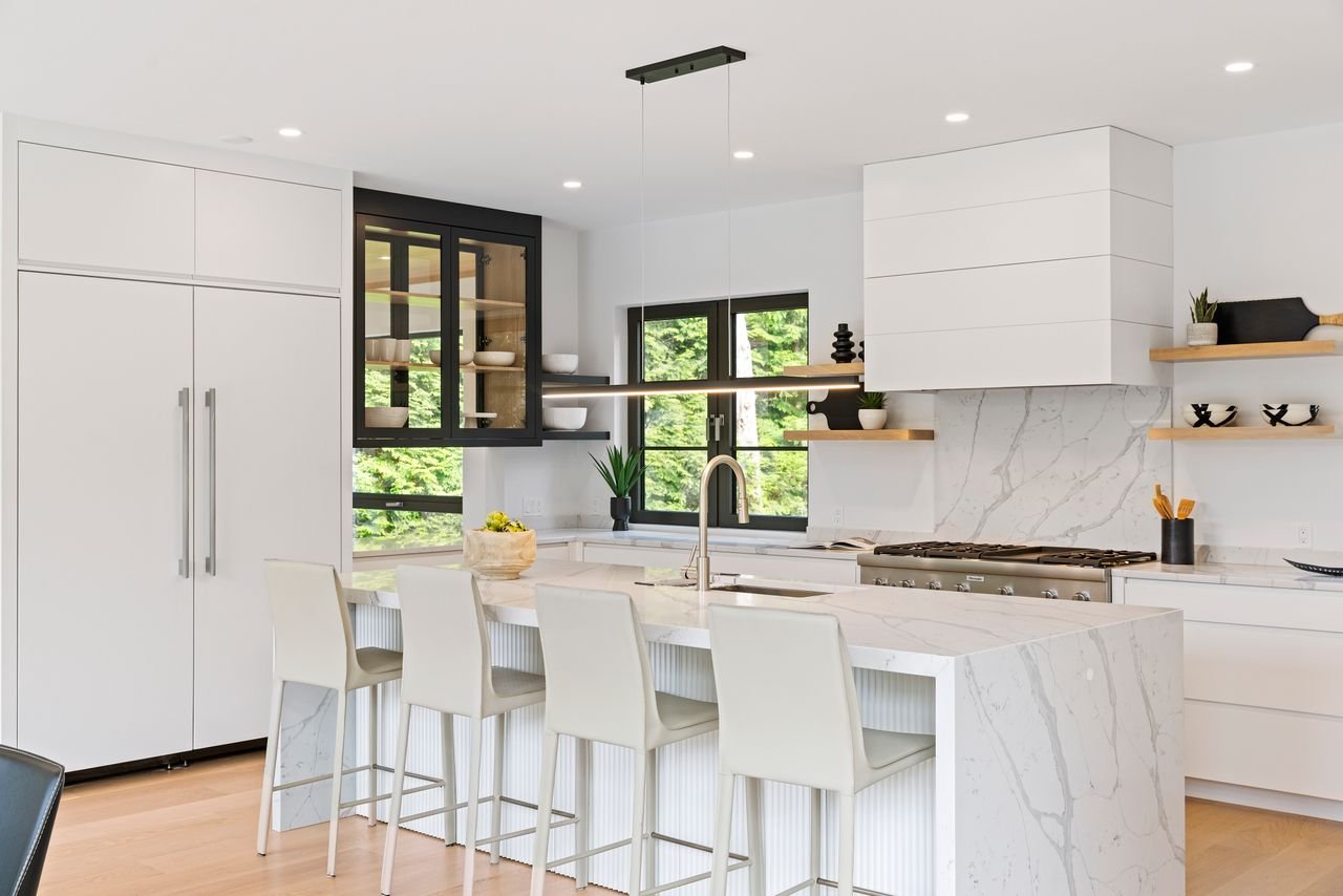 Modern white kitchen with marble island, four cream barstools, black window frames, open shelving with decor, and a large window showing greenery outside.