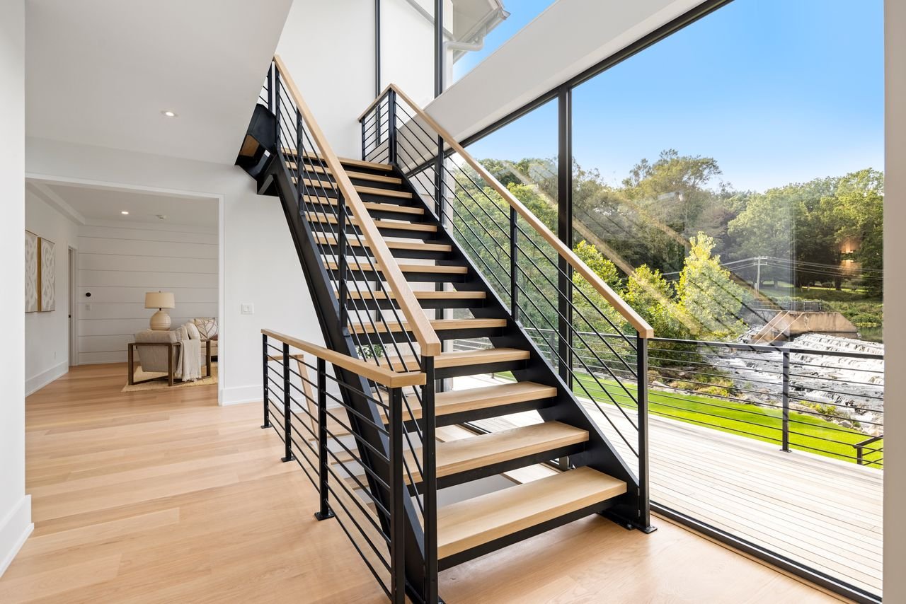 Interior view of a modern home with a staircase featuring black metal railing, wooden steps, and a large floor-to-ceiling window overlooking a lush green landscape and a waterfall.