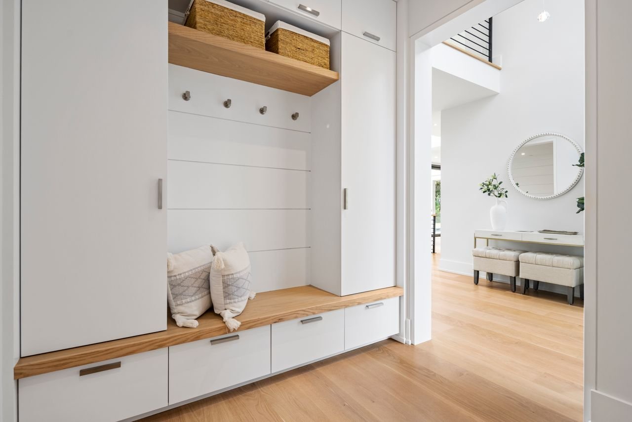Interior view of a modern home entryway with white built-in storage, wooden accents, and light wood flooring leading into a bright living space with a mirror and a white console table.