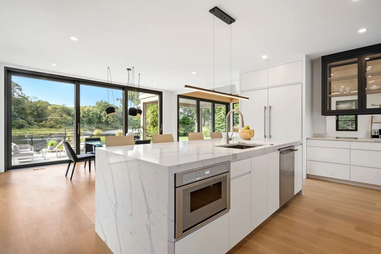 Modern open-concept kitchen with a white marble island, light wood flooring, and large floor-to-ceiling windows overlooking a scenic outdoor view.