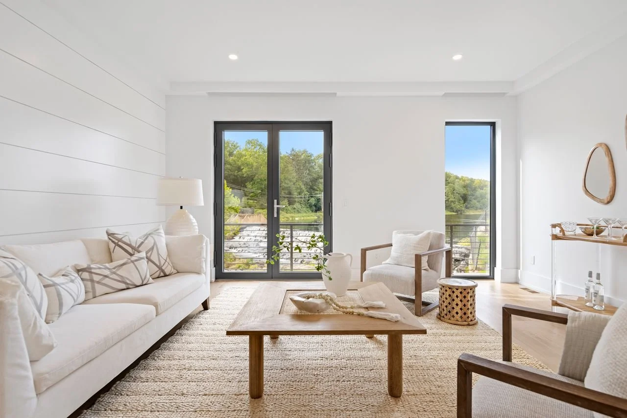 Bright living room with white sofa, wooden coffee table, armchair, and large windows with a view of green trees and a river.