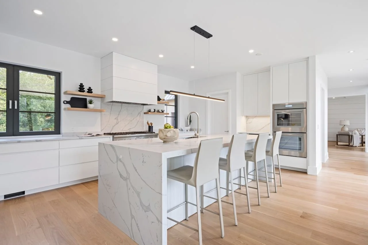 Modern white kitchen with an island, barstools, open shelves, and built-in appliances, with natural light coming through a window and hardwood floors.