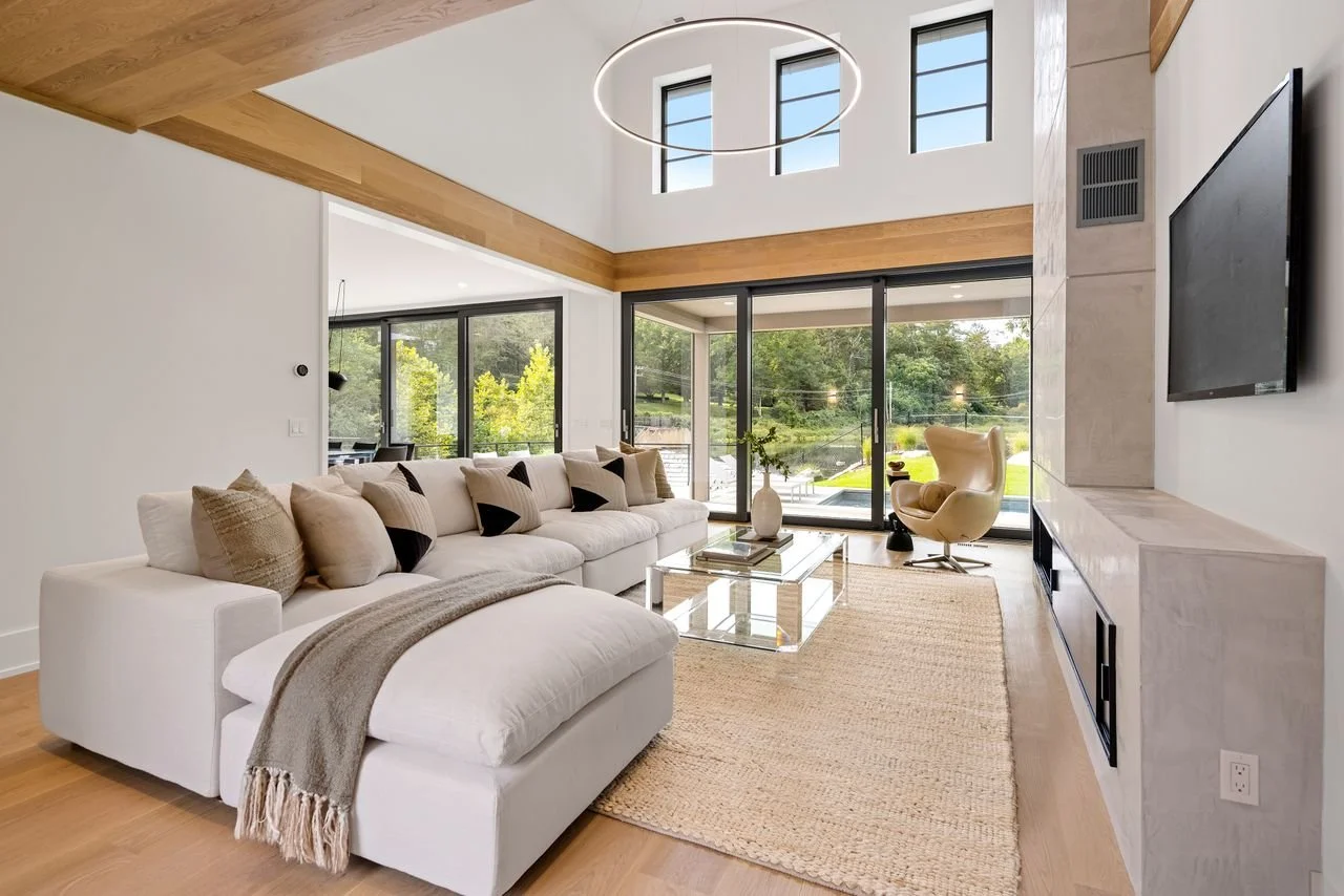 Modern living room with large white sectional sofa, glass coffee table, beige area rug, and a high ceiling with large windows and circular chandelier.