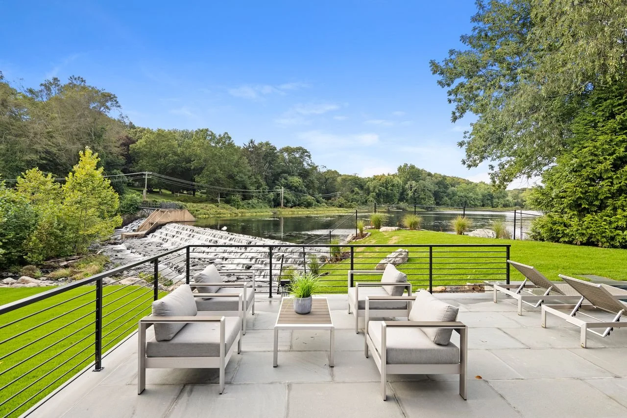 Outdoor patio with gray cushioned chairs, a small table with a plant, and sun loungers overlooking a river with a small waterfall, trees, and a clear blue sky.